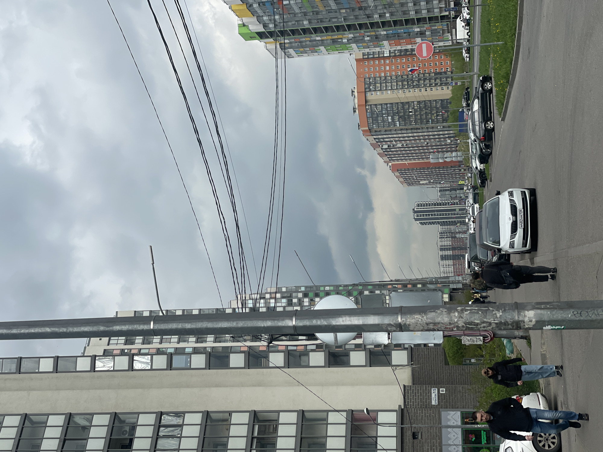 The image depicts an urban scene in an unidentified city. Tall apartment buildings of varying heights and colors line the background, indicating a densely populated area. A street with parked cars and pedestrians is visible on the right side of the image. A utility pole with power lines runs across the foreground, partially obstructing the view of the buildings. Three pedestrians are walking on the sidewalk next to the street. The sky is overcast, suggesting a cloudy or possibly rainy day.