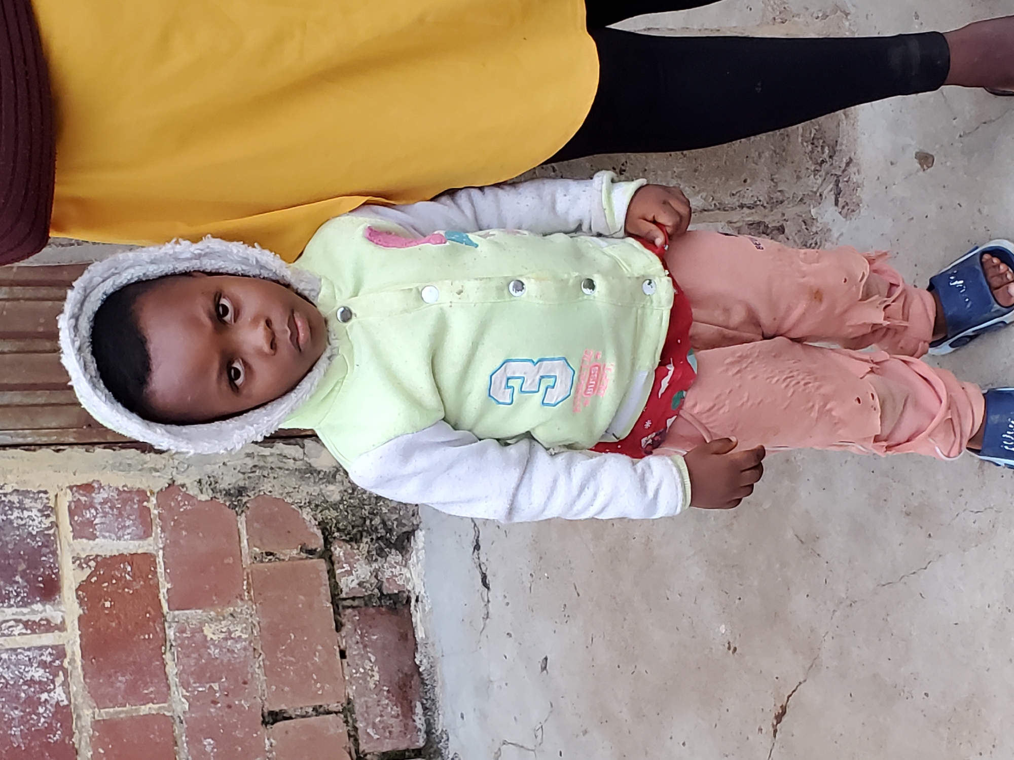 A young child stands in front of a brick wall and a person wearing a yellow garment. The child is wearing a light green hooded jacket with white sleeves, a red undershirt, and torn pink pants. The child also wears blue sandals. The ground is concrete with cracks. The image appears to be taken in an outdoor urban environment.
