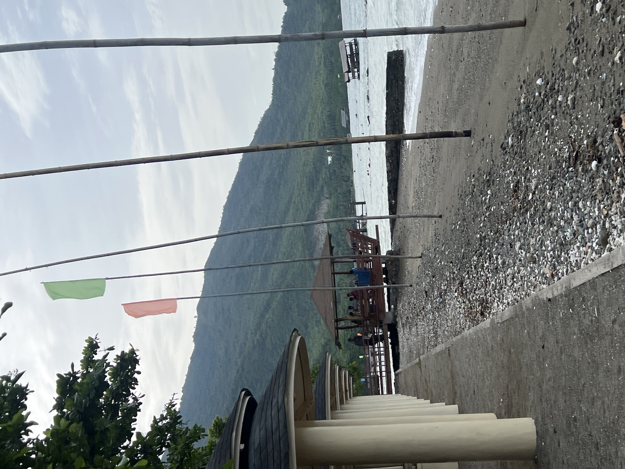 The image shows a coastal scene in Banaybanay, Philippines. A beach with light-colored sand and darker, pebbly areas is visible. The water is choppy with small waves. In the distance, a large, lush green mountain dominates the background. Several bamboo poles are suspended horizontally in the foreground, likely part of a structure or decoration. Two flags, one green and one salmon-colored, are attached to the poles. A wooden structure with people visible on it extends over the water. A row of small, round, roofed structures lines the foreground. A concrete walkway or wall runs along the edge of the beach. Some vegetation is visible on the left side of the frame. The sky is overcast.