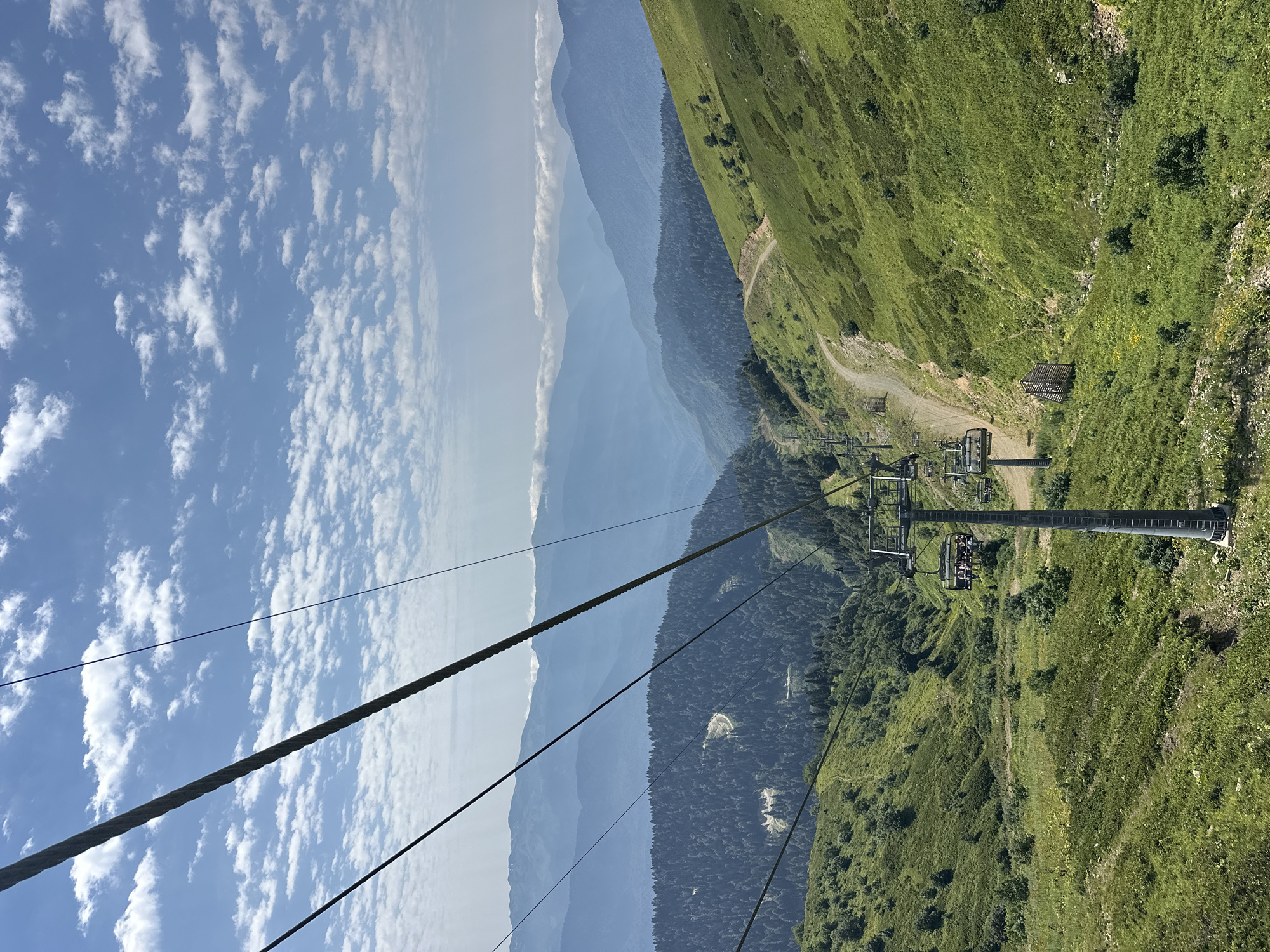 The image captures a view from a ski lift ascending a lush green mountainside in Estosadok, Russia. The ski lift’s cable is prominent in the foreground, stretching diagonally across the frame. Below, the ski lift chairs, some occupied by passengers, descend down the slope. The mountainside is covered in vibrant green grass and scattered trees, with a winding dirt path visible on the right. In the distance, layers of progressively bluer mountains recede into the hazy horizon under a bright blue sky dotted with fluffy white clouds. The overall scene depicts a scenic mountain landscape on a sunny day.