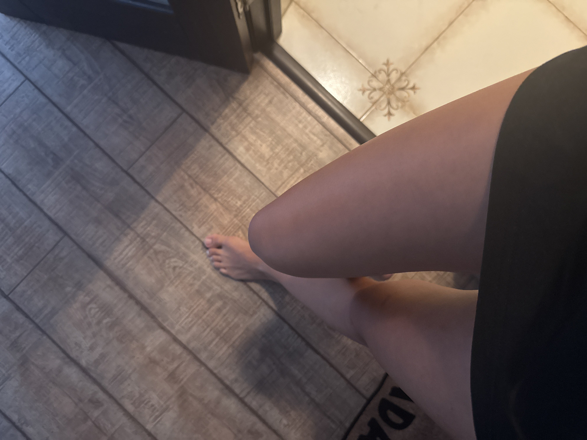 The image shows a pair of bare legs and feet resting on a wooden floor. The legs are smooth and appear to be from a young adult. The feet are positioned such that the toes are visible. The wooden floor is laid in planks, arranged diagonally across the frame. A dark mat with some lettering is partially visible on the left side of the floor. To the right of the wooden floor, there is a tiled floor with a decorative pattern. The overall lighting suggests an indoor setting, possibly a home. There are no discernible elements that would indicate a specific location within Chișinău, Moldova.