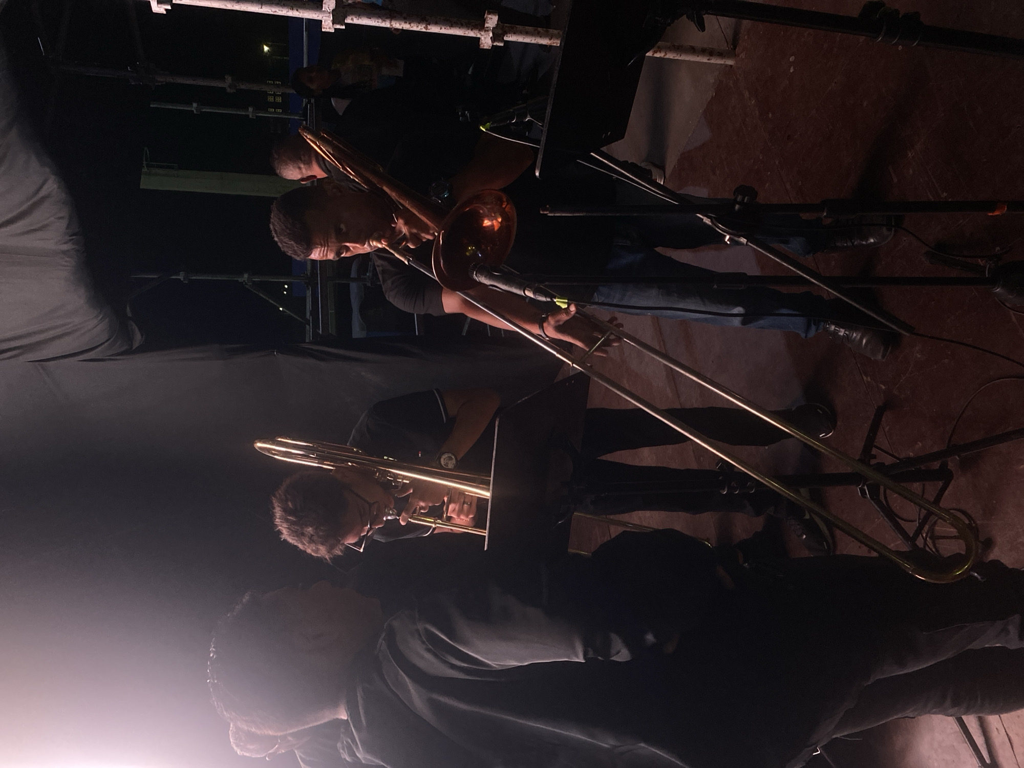 The image shows a band playing trombones on a stage. Three people are visible playing trombones. One is wearing glasses. Another is wearing a watch. The stage is dark, with some equipment visible. The location is Tinaquillo, Venezuela.