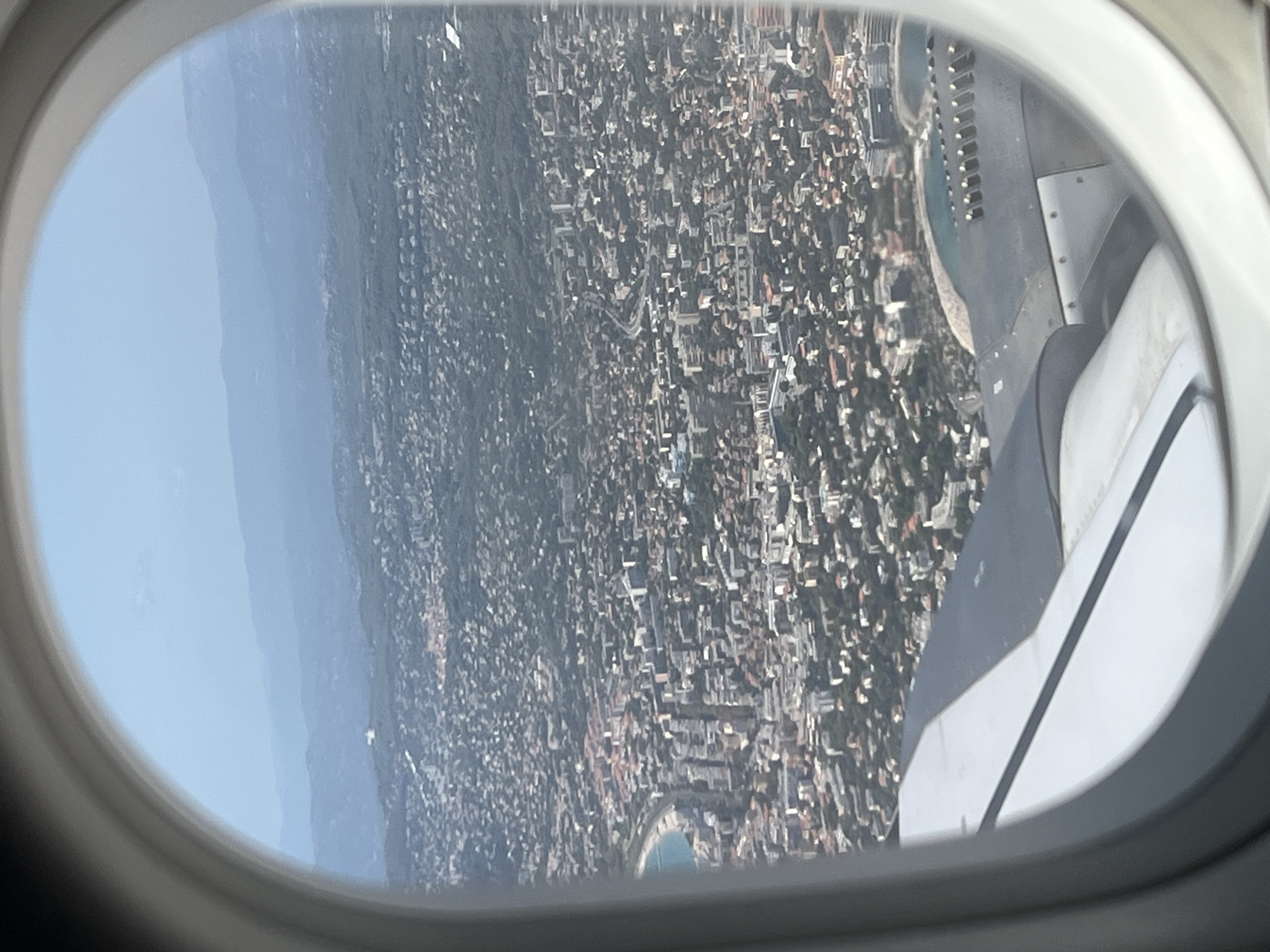 This aerial view, captured through an airplane window, looks down upon the coastal city of Saint-Laurent-du-Var, France. The scene is dominated by a dense urban landscape of buildings with red-tiled roofs, interspersed with trees and greenery. In the foreground, a portion of the airplane's wing is visible, its metallic surface reflecting the sunlight. The city extends to the coastline, where a bay with turquoise water meets a sandy beach. Beyond the city, rolling hills and mountains fade into a hazy blue sky. The overall impression is one of a vibrant Mediterranean city nestled between the sea and the mountains.