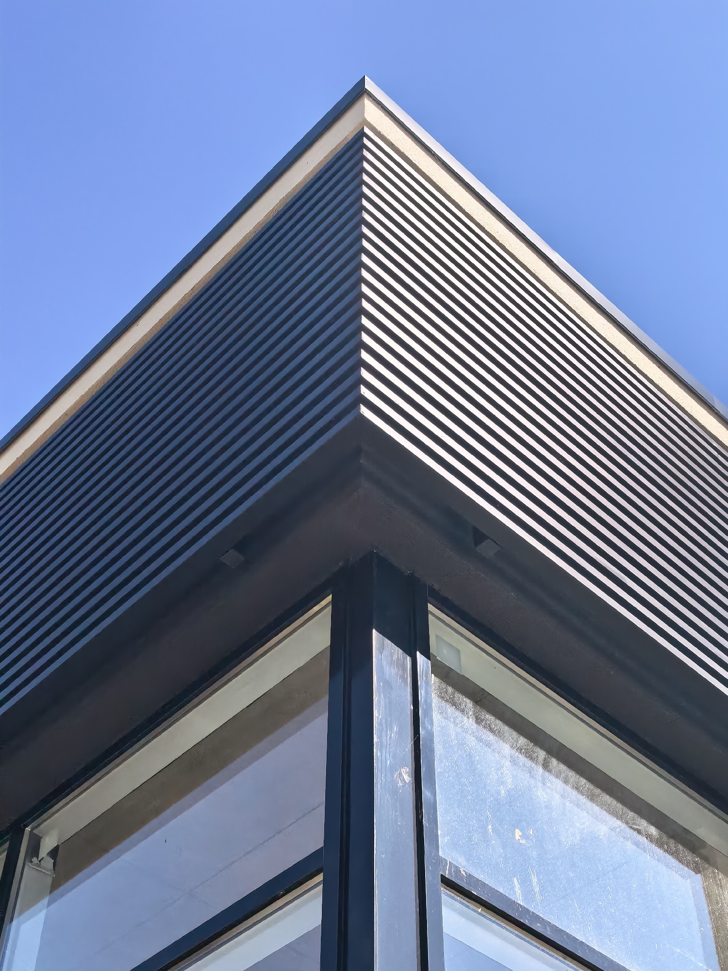 The image shows a close-up, low-angle exterior view of a modern building corner with a blue sky as the background. The building features a ribbed, dark-colored facade above a large window. The ribbed section is composed of horizontal, dark gray slats with bright, thin gaps between them, creating a textured effect. A lighter, beige band runs along the top edge of the ribbed facade. The window below is framed by thick, dark metal mullions and appears to be somewhat dirty, reflecting the sky and possibly some internal lights. The overall impression is of clean, contemporary architectural design. The specific location of this building in Sestroretsk, Russia, is not visually discernible from the image itself.
