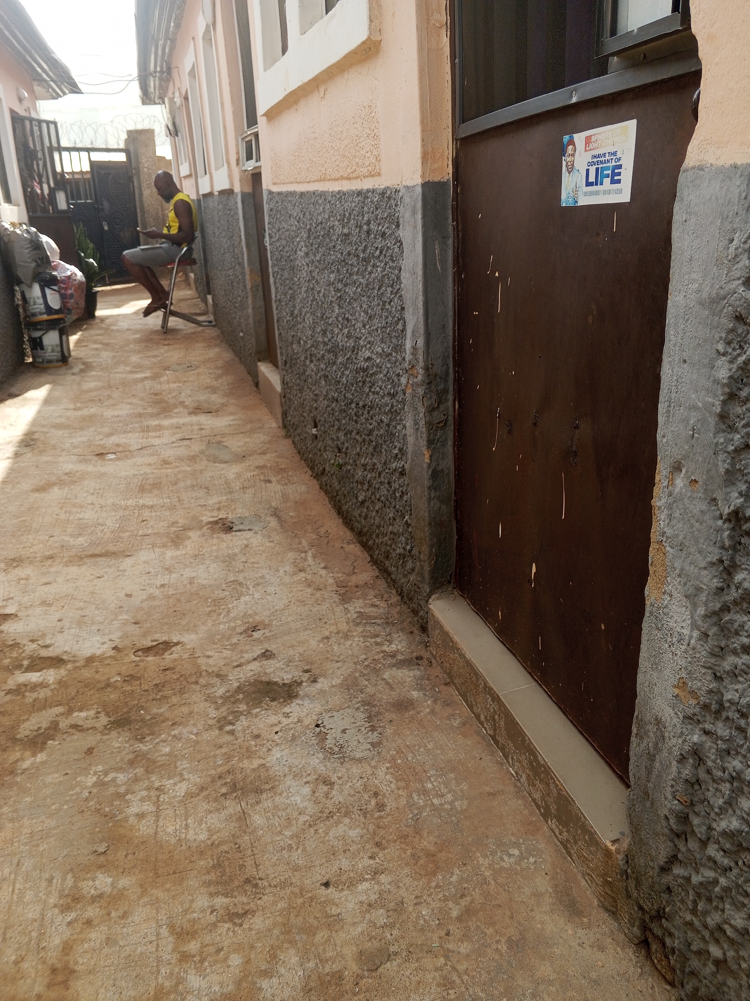 A narrow alleyway in Abuja, Nigeria, is depicted. A man is sitting on a small stool at the end of the alleyway, facing away from the viewer. The alleyway is between two buildings, one with a brown door that has a sticker that says "I Have the Covenant of LIFE" on it. The buildings appear to be residential. The ground is cracked and uneven. Some discarded objects are stacked near the man. There is a fence in the background, topped with barbed wire.