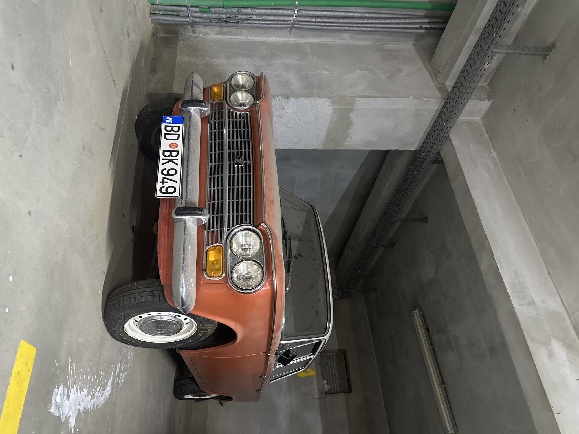 An aged, faded orange sedan, identified by its license plate as BD BK 949, is parked in an indoor concrete garage. The car, a vintage model, exhibits signs of wear and tear, with a dusty windshield and visible rust on its body. Its chrome bumper and grille are intact, and it rests on white-rimmed wheels.
The garage environment is utilitarian and dimly lit, with exposed concrete walls and ceiling. Gray metal conduits and pipes run along the ceiling and walls, suggesting an industrial or service area. Green pipes are also visible on the left side of the frame. The floor is concrete, marked with a yellow line and splatters of white fluid, potentially paint or a cleaning agent. The car is positioned such that its front is angled slightly towards the viewer. The background shows more concrete structures and a ventilation grate on the right wall. The overall impression is of an old car stored in a functional, somewhat neglected, urban infrastructure setting in Budva, Montenegro.