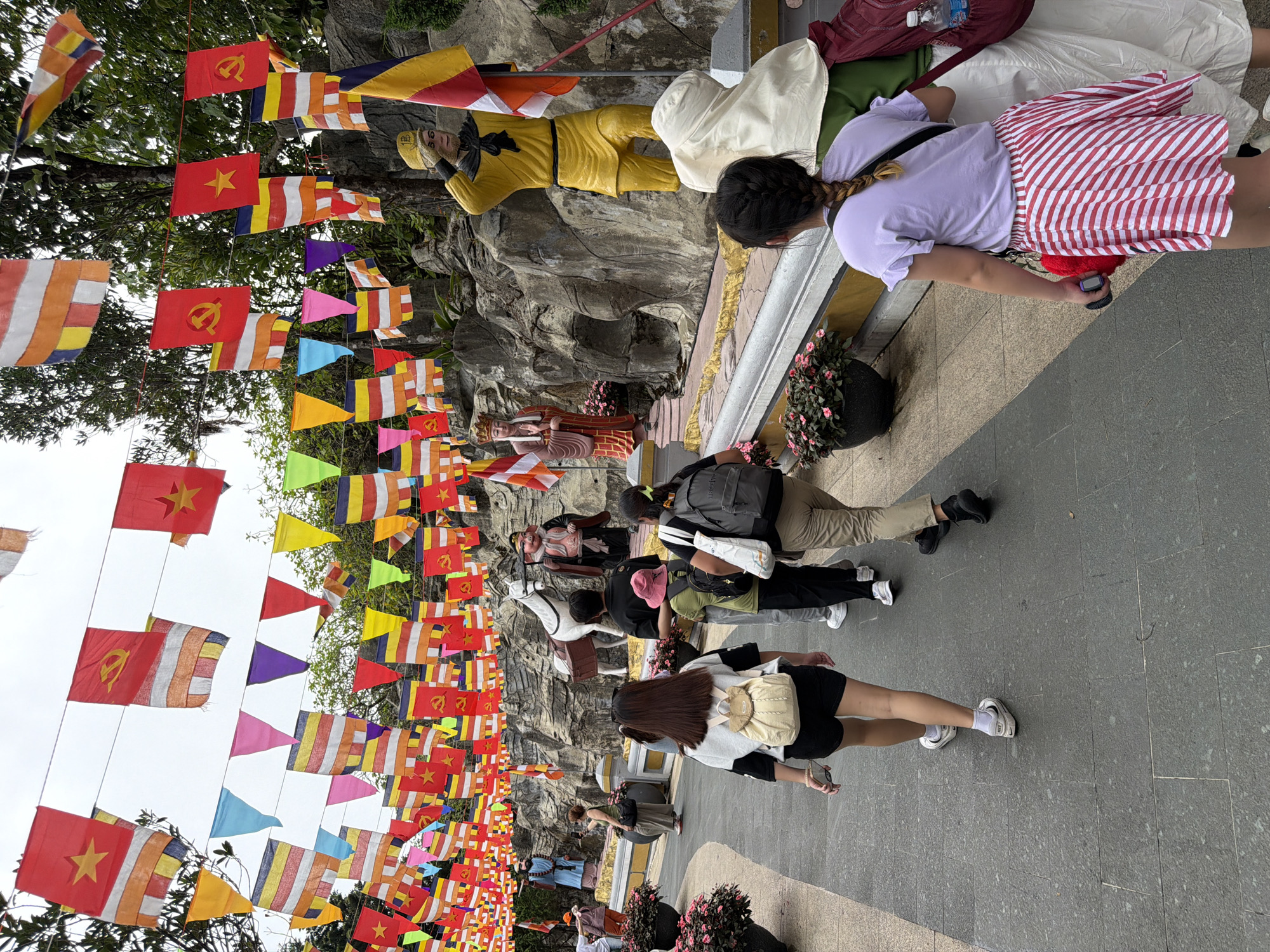 The image captures a vibrant scene at a temple complex in Đà Nẵng, Vietnam, possibly within the Sơn Trà Peninsula. The foreground is dominated by a paved walkway where several individuals are walking. A woman in the center, wearing black shorts and a cream-colored backpack, walks away from the camera. To her right, a person in a black shirt and a pink hat is also walking away. Further ahead and to the right, a woman in a light purple t-shirt and a red and white striped skirt is seen from the back.
The walkway leads towards a series of stone sculptures and structures, likely part of the temple's artistic display. A prominent sculpture of a figure in a yellow uniform, saluting, stands on the right. To the left of this figure is a white horse sculpture. Behind these sculptures, intricate rock formations are visible.
The scene is further enhanced by a multitude of colorful flags and pennants strung across the pathway overhead. These flags, predominantly red and yellow, with some also featuring blue and white stripes, create a festive and spiritual atmosphere. In the background, lush green trees suggest the natural surroundings of the temple complex. The overcast sky indicates a potentially humid day. The overall impression is one of a cultural and religious site teeming with visitors.