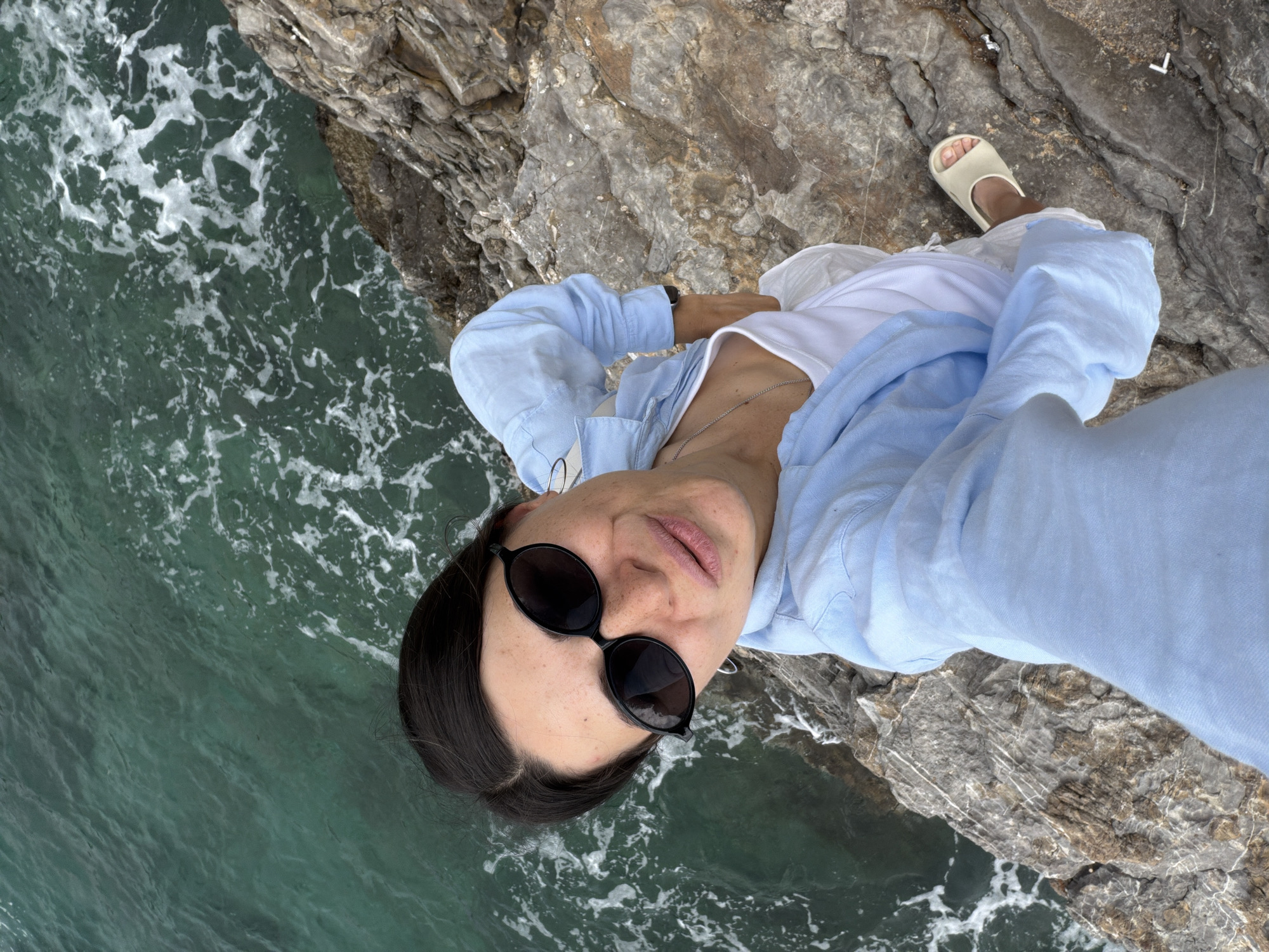 A selfie taken from above shows a woman with dark hair and sunglasses standing on a rocky cliff overlooking the sea. She is wearing a white t-shirt and a light blue shirt. The sea is turquoise, with whitecaps near the rocks. The rock formation is grey with various textures. A single sandal is visible at the edge of the cliff. This image was likely taken in Budva, Montenegro, given the coastal landscape.