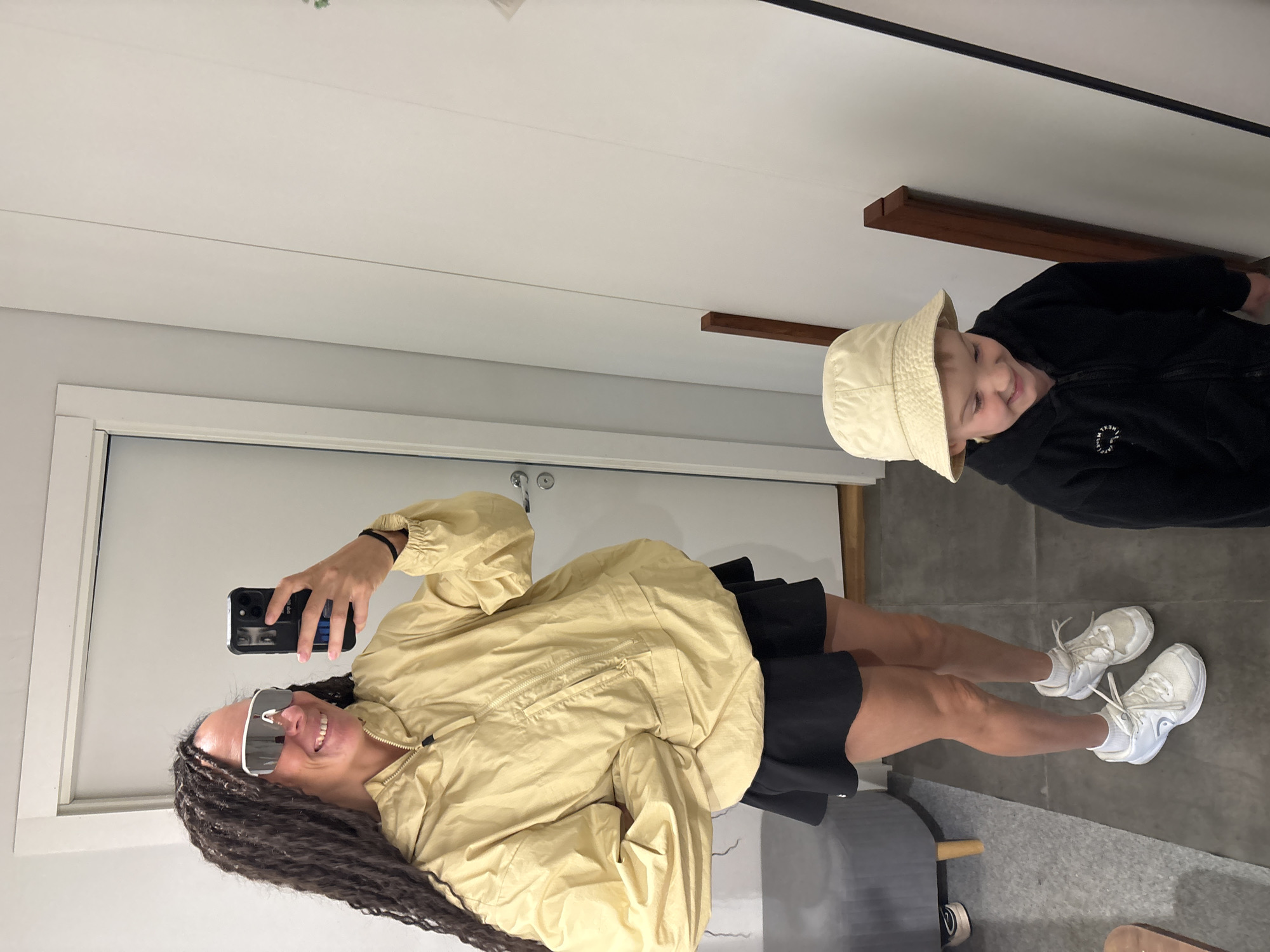 In a room with white walls and a grey floor, a woman with long dark braided hair, wearing a yellow hooded jacket, a black skirt, white sunglasses, and white sneakers, takes a mirror selfie holding a black smartphone. To her right, a young boy wearing a beige bucket hat and a black hooded jacket smiles down. The room appears to be a fitting room, with a gray ottoman visible to the left.