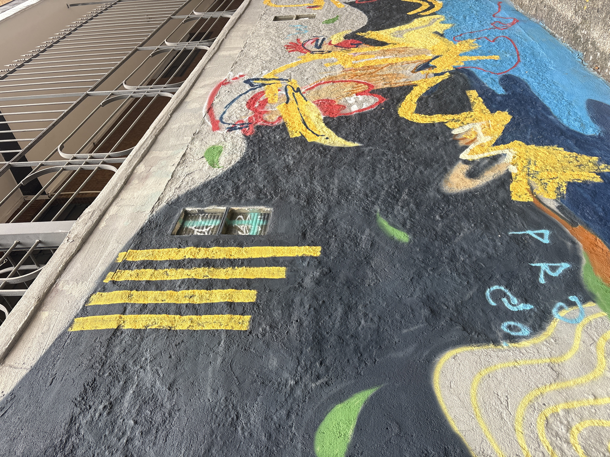 The image shows a close-up view of a vibrant mural painted on a textured, dark gray stucco wall in São Paulo, Brazil. The mural features several stylized rooster figures with bright yellow bodies, red combs, and blue outlines. In the upper left portion of the wall, there are four vertical yellow stripes. A small, barred window with green bars is visible between the stripes and the roosters. The wall is situated beneath a concrete overhang with a metal railing, suggesting it's part of an exterior wall of a building, possibly a balcony or second story. The lower part of the mural transitions to bright blue and yellow abstract shapes, with hints of orange and green. A portion of what appears to be a light-colored, abstract form with yellow curves is visible on the far left. The overall impression is one of urban street art.