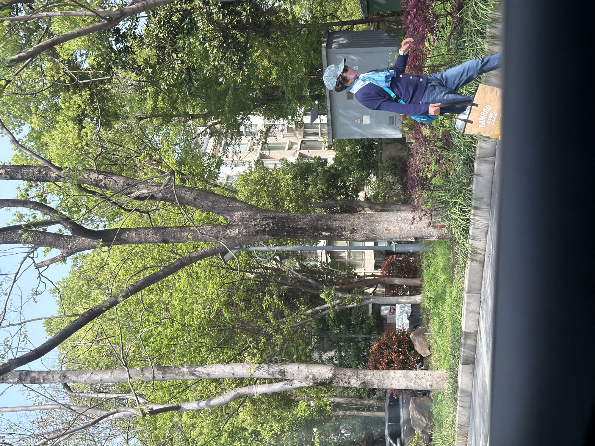 A man is walking along a sidewalk in Yuhuatai District, China, carrying a brown bag that reads "BAMBOO IS COOL." He wears a blue jacket, jeans, a light blue scarf, and a blue patterned cap. The scene includes trees, grass, bushes, and a building in the background. A utility box is also visible near the man. The image appears to be taken from inside a vehicle.