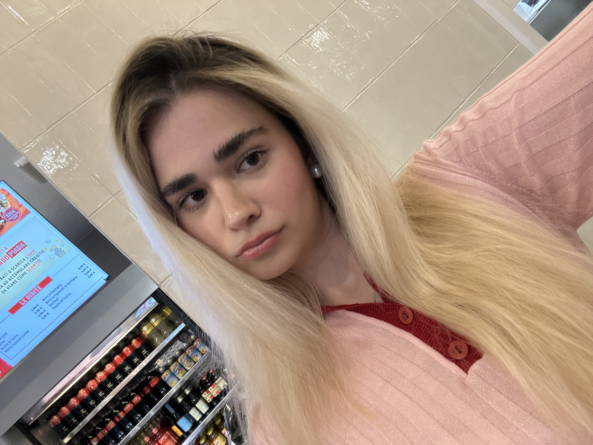 The image is a medium close-up selfie or portrait of a young woman with long, straight, blonde hair, looking directly at the camera with a neutral, slightly contemplative expression. She has dark, defined eyebrows and is wearing a light pink, ribbed long-sleeved top with a reddish-pink collar and two matching buttons. A single pearl earring is visible on her right ear.
The setting is an indoor commercial establishment, likely an Italian casual eatery, possibly a piadineria, in Milan, Italy. The background features light-colored, glossy tiled walls. To the left, a digital menu board in Italian displays various food and drink options. Prominently featured is "PIADA" and a section titled "LE BIBITE" (The Drinks). Listed drink options include "Birre in bottiglia" (bottled beers) at 3.50€ and 3.90€, "Birre artigianali in bottiglia" (craft bottled beers) at 4.50€, "Red Bull" at 3.50€, and "Succhi di frutta" (fruit juices) at 2.90€. Also mentioned are "Bibite in vetro" (bottled soft drinks) at 3.00€ and "Bibite speciali Lurisia" at 3.20€. The board encourages customers to "SCARICA L'APP" (download the app) for "SCONTO!" (discount!). Below the menu board, a refrigerated display case is packed with a variety of bottled beverages, including Coca-Cola, other sodas, and beers, with the brand "Ichno Khnus" visible on some labels. The indoor lighting suggests an indeterminate time of day.
