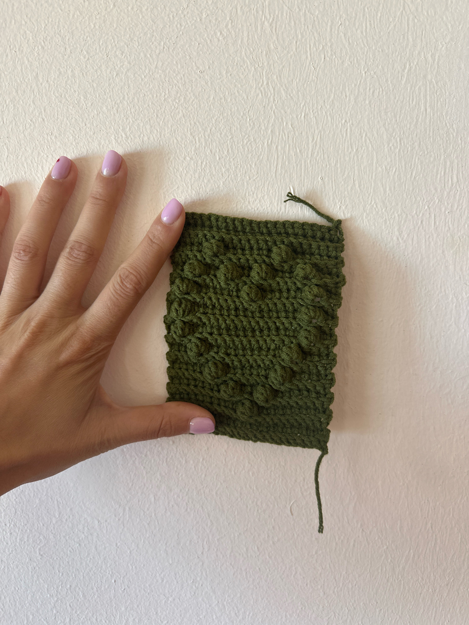 A hand with light pink nail polish is holding a square swatch of green crochet fabric. The fabric has a raised, textured pattern resembling small bumps. The swatch is positioned on a white, textured wall. A loose green yarn tail extends from the bottom right corner of the swatch. The scene is illuminated with natural light.