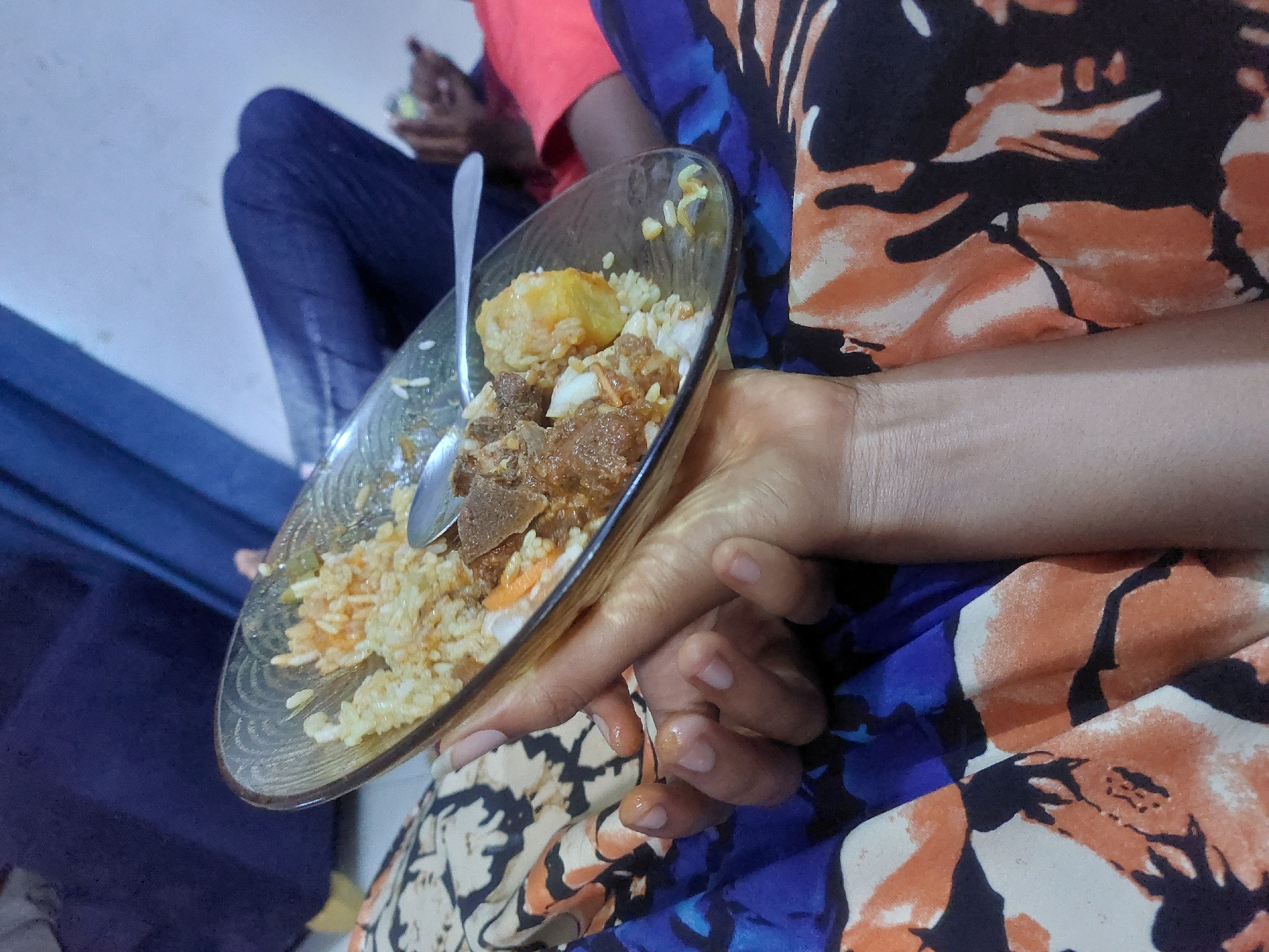 Here's a detailed description of the image:
A person is holding a clear glass plate filled with a meal consisting of rice, meat, and a yellow, cubed vegetable (possibly potato or squash). The person is holding a spoon on the plate. The person is wearing a floral-patterned garment in shades of blue, orange, and white. The background shows another person wearing red, seated in a blue chair. Given the food type, setting and clothes and other hints of the image, this image is taken in a typical home in Dar es-Salaam, Tanzania.