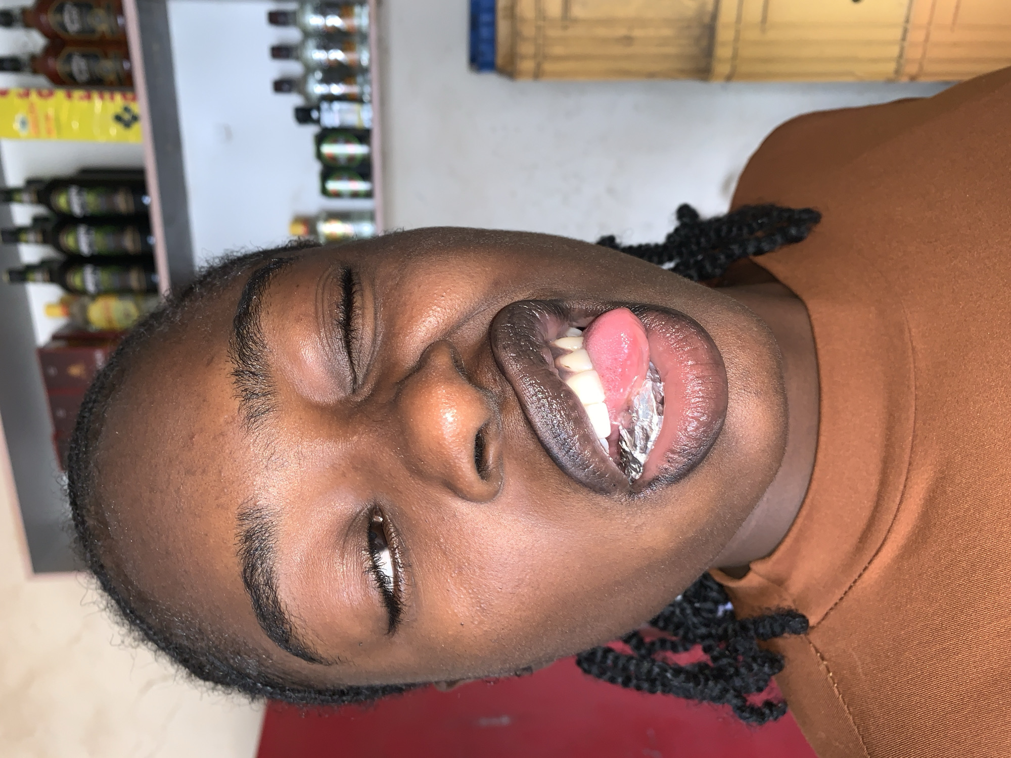 The image displays a young Black female, captured indoors in New Stadium, Nigeria. She is positioned centrally, wearing a plain, high-neck, rust-brown top. Her dark hair is styled in small braids that frame her face. The subject's face is illuminated, highlighting her forehead and nose. She presents a deliberate expression: her right eye is fully closed in a wink, while her left eye is partially open and gazes to the viewer's right. Her mouth is wide open, with a pink tongue extended forward. A crumpled piece of metallic material, possibly aluminum foil, rests on her lower front teeth, pressing down on the tongue. Her lips appear to be coated with a dark substance.
The background is split between two wall colors. To the left, a solid red wall is visible. To the right, the wall is a lighter, off-white or cream color. Two white shelves are mounted on this lighter wall. The upper shelf holds multiple dark-colored bottles, possibly alcoholic beverages, and a bright yellow rectangular package. The lower shelf contains several clear bottles, green beverage cans, and one dark bottle. In the lower right portion of the frame, a segment of a textured, yellow-brown object, resembling a large container or piece of furniture, is visible.
