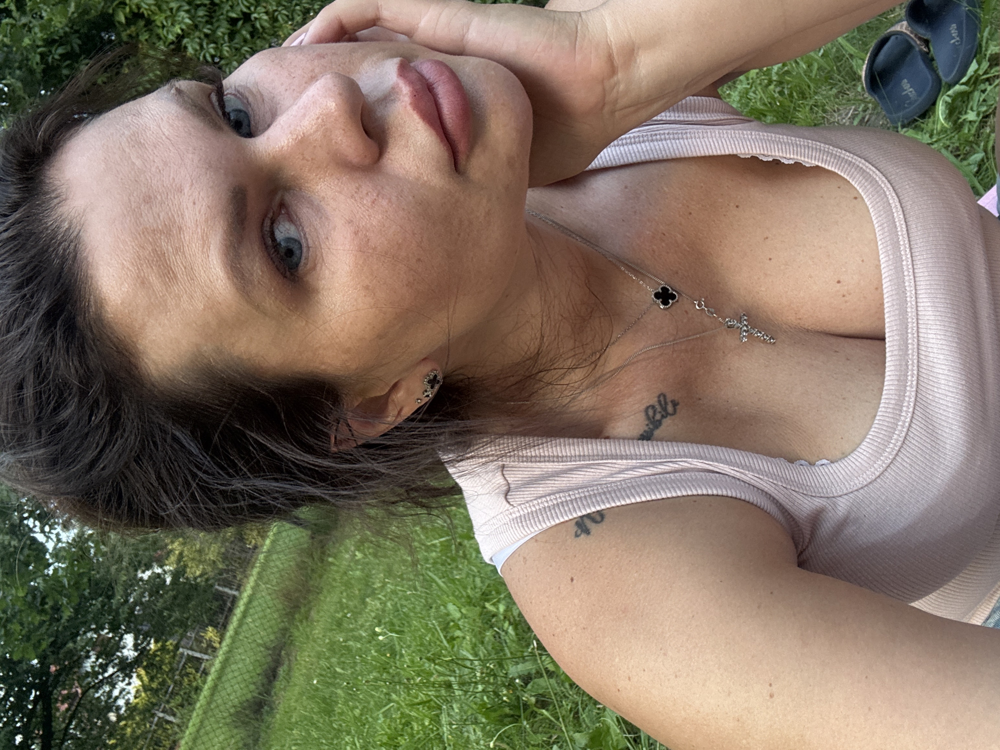 A woman is captured in a close-up, outdoor portrait, likely in a park or grassy area in Katowice, Poland, given the context. She is wearing a light pink ribbed tank top, which reveals her cleavage and a cross necklace with a clover charm. Tattoos are visible on her upper arm and chest. Her right hand is resting on her cheek, and she is looking towards the camera with blue eyes and wearing subtle makeup. The background consists of green grass, trees, and a fence.