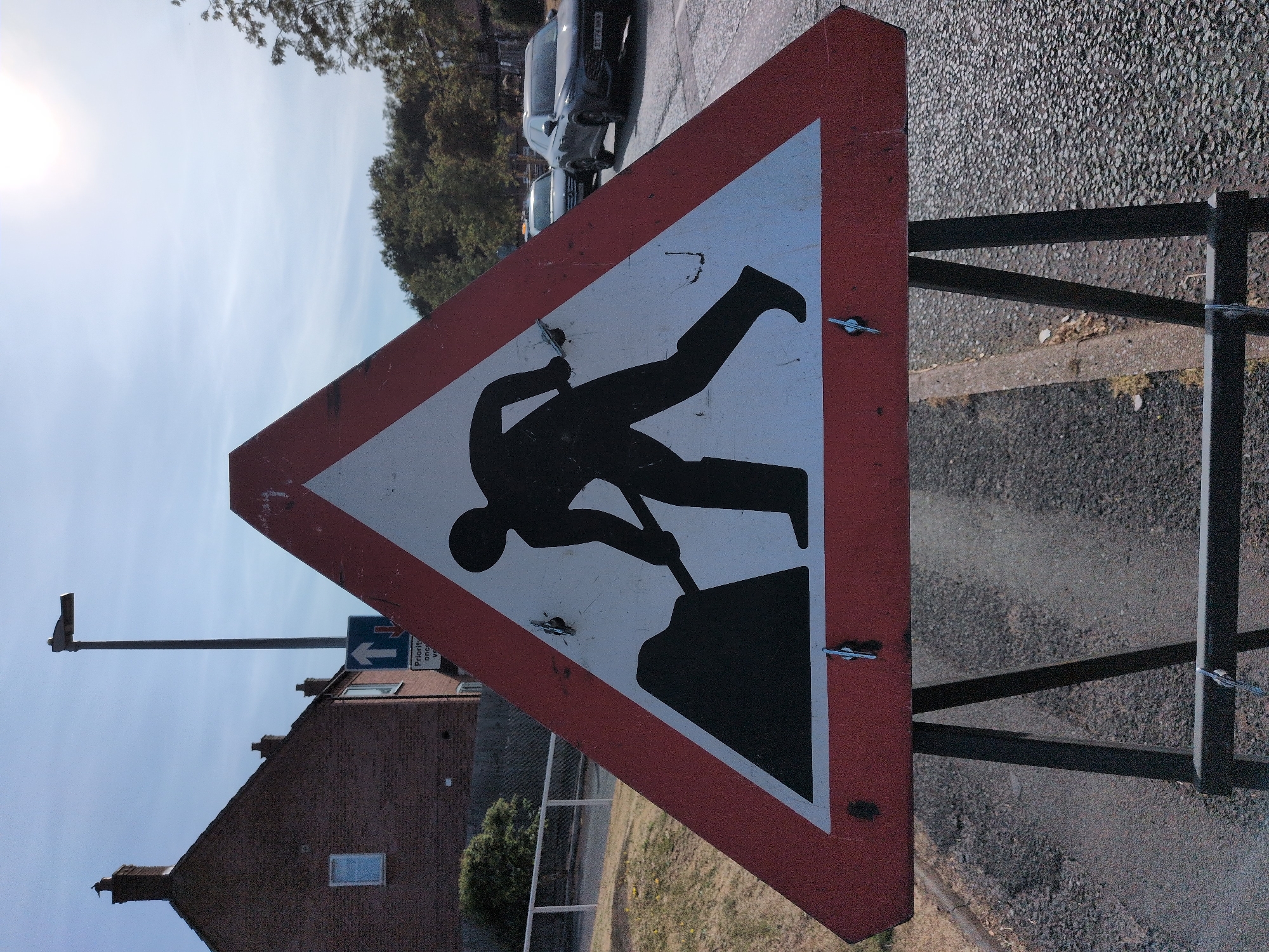 A roadworks warning sign, depicting a silhouette of a person digging with a shovel, is erected on a metal A-frame stand. The sign is triangular with a red border and a white background, featuring the black silhouette against the white. The sign is placed on a road surface, with a concrete curb and a grassy verge visible to the left. In the background, a brick building with a pitched roof and a chimney stands, accompanied by a lamppost. Further in the distance, trees and two cars are present. The sky is visible overhead with the sun shining brightly. The location is identified as Dawley, United Kingdom.