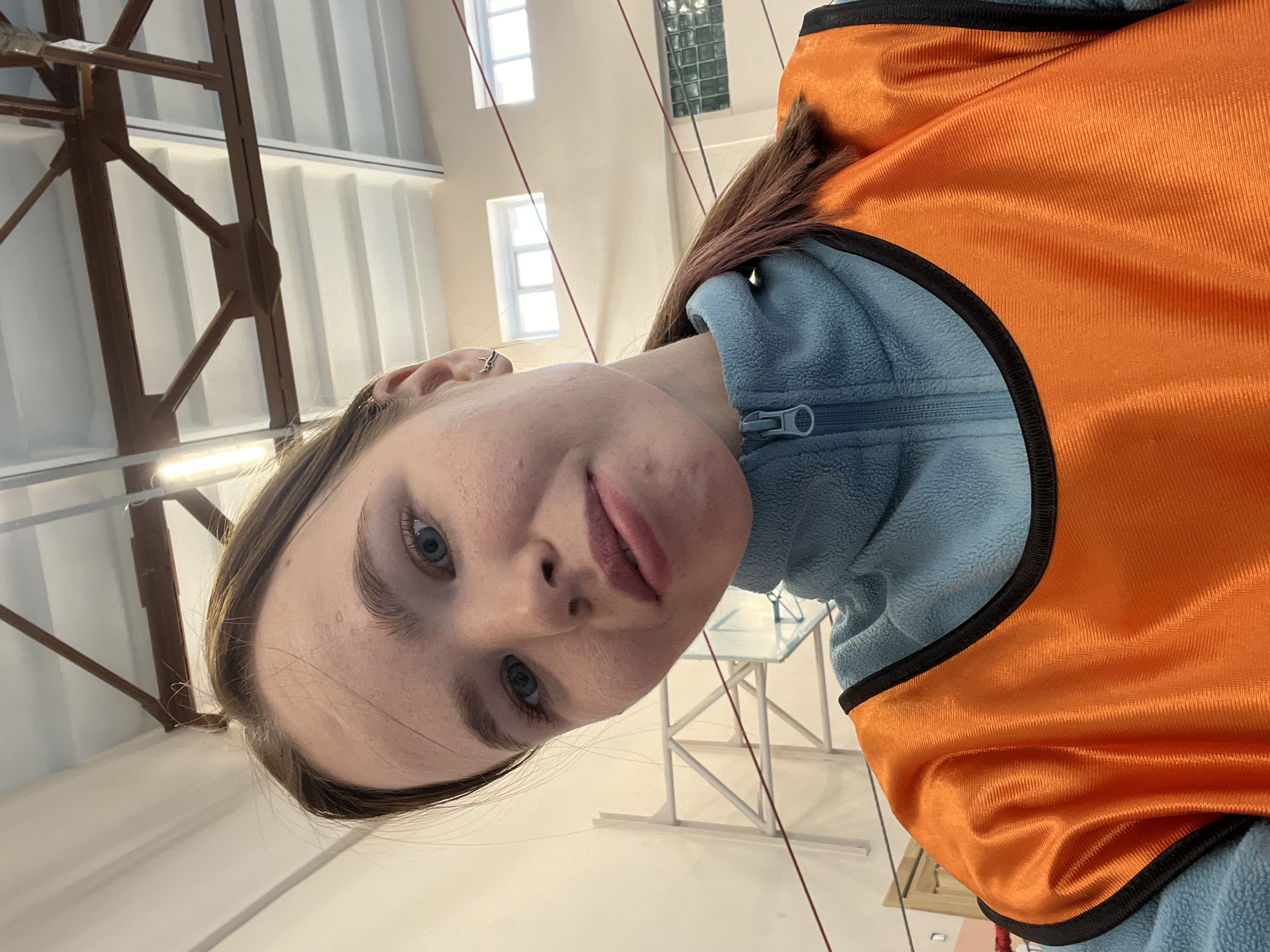 The image is a selfie of a young woman. She has fair skin, blue eyes, and light brown hair with some reddish highlights. She is wearing a light blue fleece jacket, over which is an orange safety vest with black trim. She has a small ring or stud in her ear.
The background shows the interior of a building. There are white walls and a high ceiling with exposed beams. Natural light is coming in through windows. There are some thin ropes hanging from the ceiling. A white metal table with a glass top is visible in the background.
The image could be taken in any interior space with similar architectural features. The location of Berezniki, Russia is only a guess as nothing indicates this location.