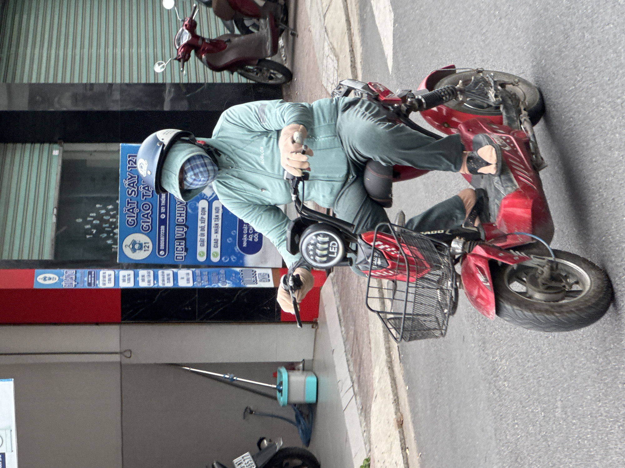A person is riding a red electric scooter down a street in Nha Trang, Vietnam. The rider is wearing a light green long-sleeved jacket, dark green pants, and a blue and white checkered face mask and hood. The scooter has a black basket on the front and the rider's hands are on the handlebars. In the background, there is a red and blue sign with Vietnamese text and images of clothes. To the right of the sign, there is a red motorcycle parked. The street is paved with asphalt and there are buildings on either side.