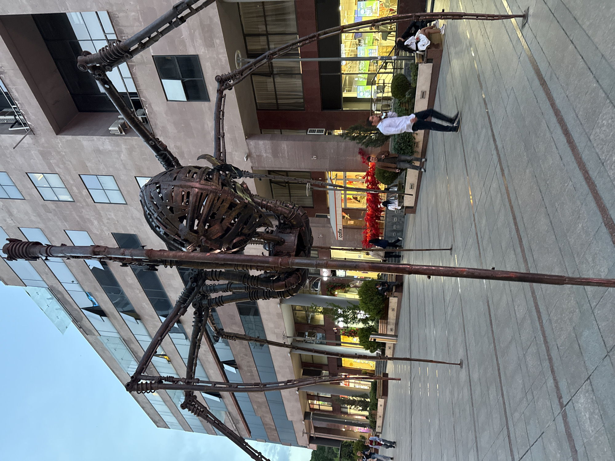 This photograph shows a large, outdoor metal sculpture of a spider in a public square. The sculpture dominates the left side of the image, with a multi-story building rising behind it. The building has large windows and a beige facade. The sculpture's legs extend towards the foreground and background, creating depth in the image.
To the right of the sculpture and building is a paved area with several people. One man in a white shirt and dark pants is walking toward the right, away from the sculpture. There are other people in the background, some sitting on benches or walking further back in the square. Some are looking at their phones. The environment appears to be a busy urban plaza with retail spaces evident through the windows of the building.
Based on the visible architectural style and the presence of the spider sculpture (known as "Maman"), the location is likely Freedom Square in Yerevan, Armenia.