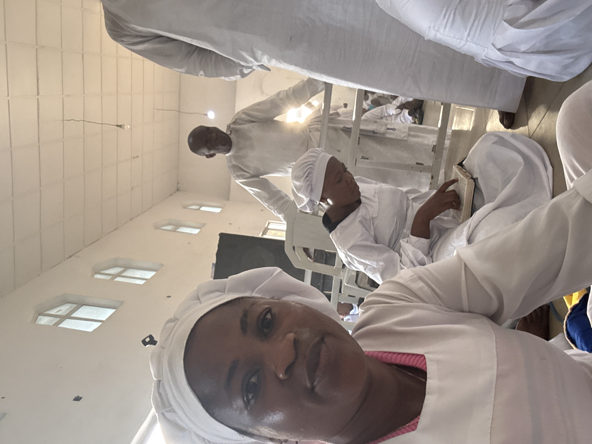 A group of people, all dressed in white robes and head coverings, are inside a building in Unknown, Unknown. The woman in the foreground appears to be taking a selfie. A man is standing in the background and a person is sitting and reading a book. The room is filled with light and there are windows along the left side of the image. There are also chairs in the background.