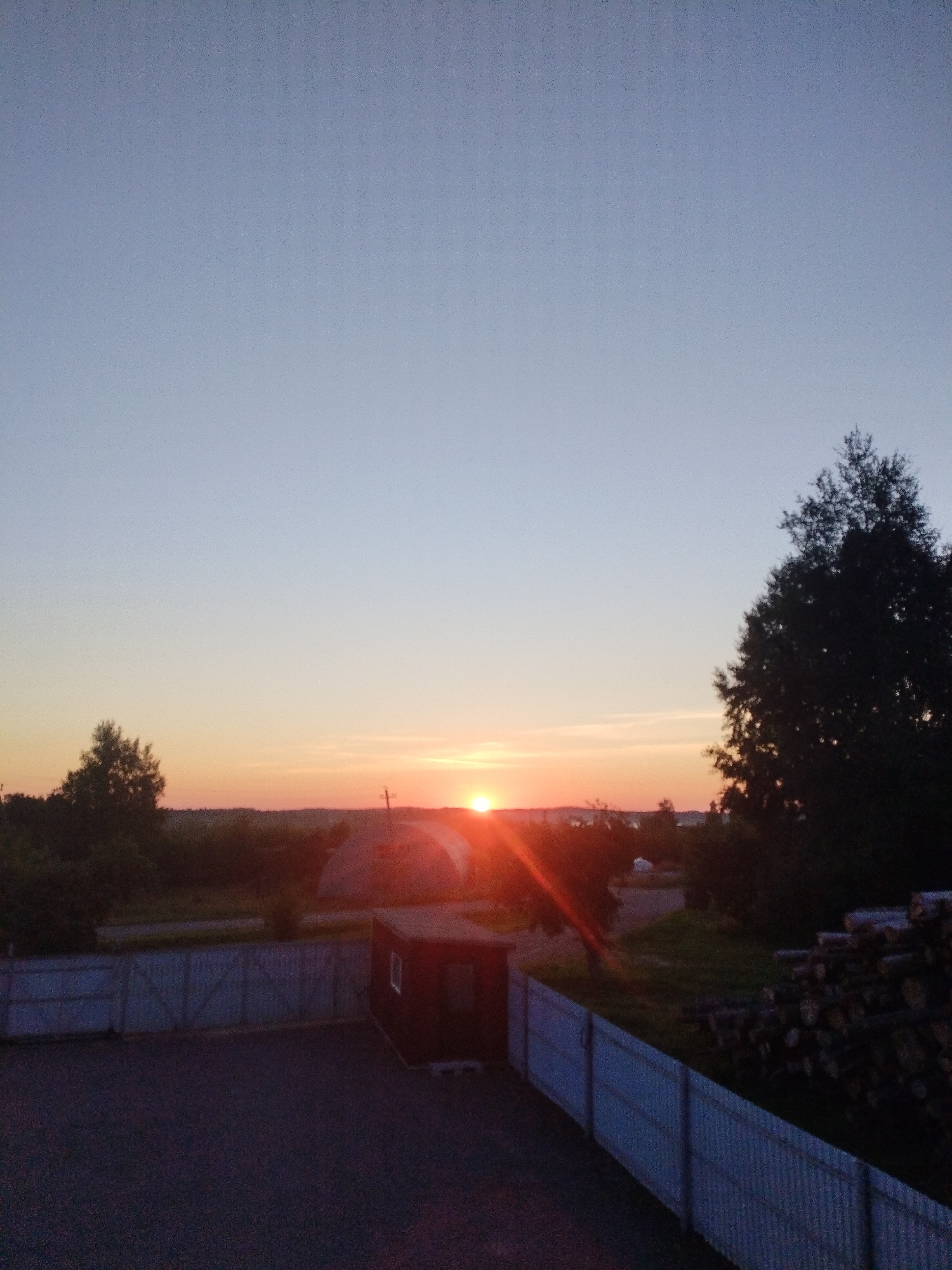 The image depicts a sunset over a rural landscape, likely in the Вароніцкі сельскі Савет, Belarus. The sky transitions from a pale blue at the top to soft orange and pink hues near the horizon, where the sun is setting. Silhouetted against the sky are trees and rolling hills. In the foreground, there is a dark asphalt surface, possibly a driveway or yard, bordered by a light-colored, corrugated metal fence. To the right of the fence, a pile of cut logs is stacked. A small, dark-colored shed with a single window is visible within the fenced area. The overall atmosphere is calm and serene.
