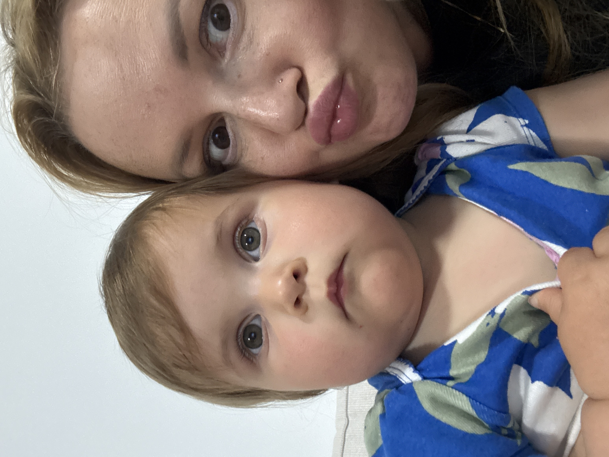The image is a rotated close-up selfie featuring an adult woman and a baby or young toddler, positioned intimately together. The adult's face occupies the upper part of the frame, showing light brown hair, warm brown eyes, and a small silver stud nose piercing. Her lips are slightly pursed, possibly with a subtle pink gloss, and she looks directly at the camera.
Below the adult, and filling the majority of the frame, is the baby. The child has light brown hair, fair skin with rosy cheeks, and large, curious light brown or hazel eyes looking straight into the camera. The baby wears a vibrant patterned outfit with blue, white, and light green abstract or camouflage-like shapes. A small hand, likely the baby's, is visible grasping the fabric of the outfit near the bottom right.
The activity captured is a casual, intimate moment between the two individuals, suggesting a parent-child bond. The lighting indicates an indoor setting. There is no visible text in the image. The scene conveys a tender and affectionate mood. The location context for this image is Ramat Gan, Israel.