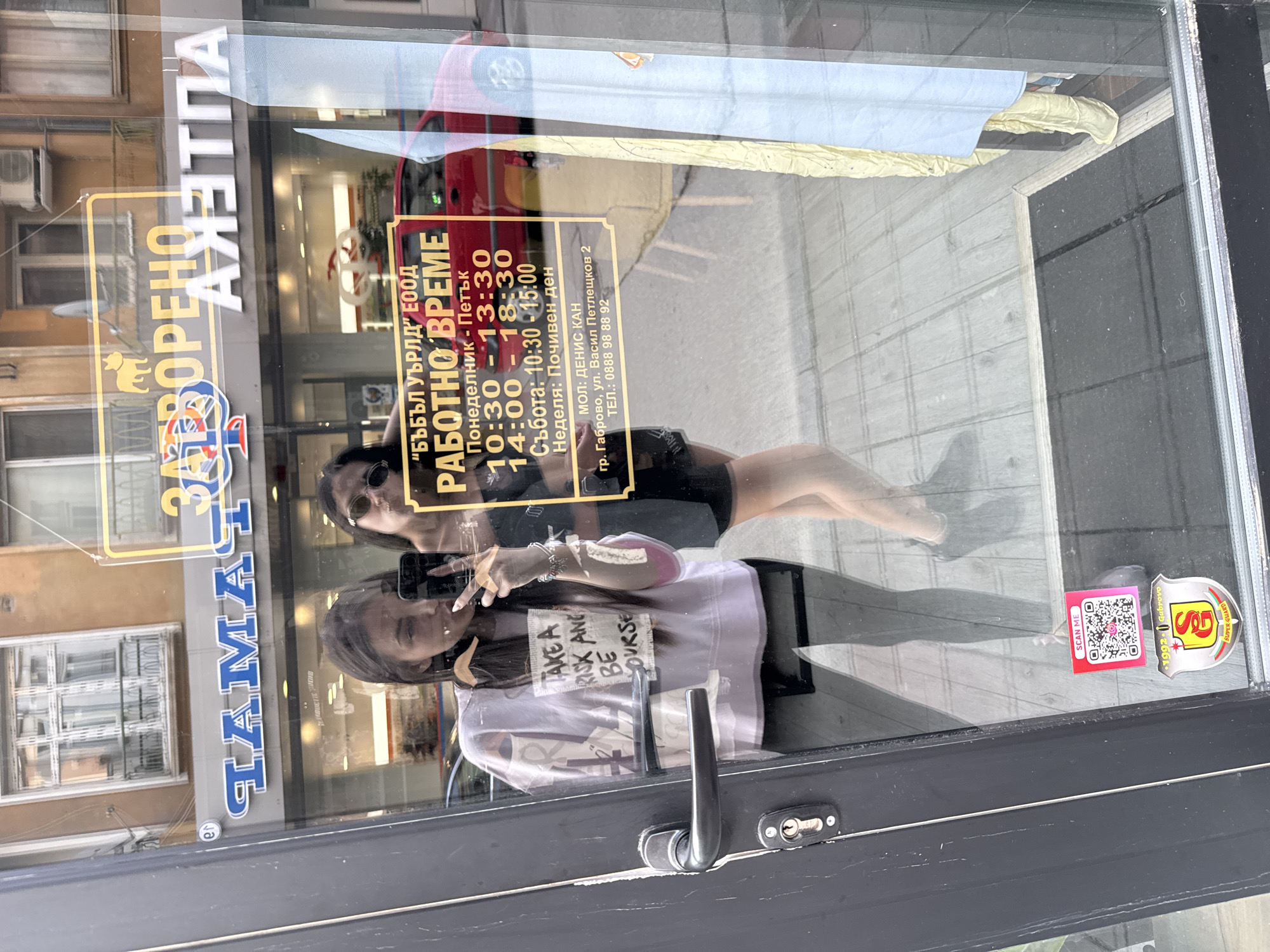The image depicts a reflective glass door and window of a business, located at Vasil Petleshkov Street 2 in Gabrovo, Bulgaria. Two young adult women are reflected in the glass, with one on the left holding a smartphone to capture a mirror selfie. This woman has long dark hair, wears a light-colored sweatshirt with the text "TAKE A RISK AND BE YOURSELF," dark leggings, and is making a peace sign with her right hand. The second woman, positioned to the right and slightly behind, is wearing sunglasses and a dark top.
Prominently displayed on the glass is a yellow sign with black text detailing the business information. The business name is "БЪБЪЛ УЪРЛД" ЕООД (Bubble World Ltd.), with "РАБОТНО ВРЕМЕ" (Working Hours) listed as Monday-Friday: 10:30-13:30 and 14:00-18:30, Saturday: 10:30-15:00, and Sunday: "Почивен ден" (Day off). Below the hours, it states "МОЛ: ДЕНИС КАН", the address "гр. Габрово, ул. Васил Петлешков 2", and a phone number "ТЕЛ.: 0888 98 88 92".
Additional elements on the glass include a pink "SCAN ME" QR code sticker and a shield-shaped sticker below it that reads "1902 Gabrovo" and "SG SUPER GUARD".
The reflections in the glass reveal an urban street scene. Across the street, a multi-story building with windows, balconies, and an air conditioning unit is visible. Reflected signs above the glass include "ЗАТВОРЕНО АПТЕКА" (CLOSED PHARMACY) with a dog logo, and another partial sign displaying blue letters that appear to be "ГАЛАНТ" or "ГАМА" reflected. A red car is parked on the street. Through the less reflective areas of the glass, a light-colored, possibly wooden or laminate, floor is visible inside the establishment, along with some draped light-colored fabric on the right. The scene is illuminated by daylight, suggesting an overcast or diffused light condition.