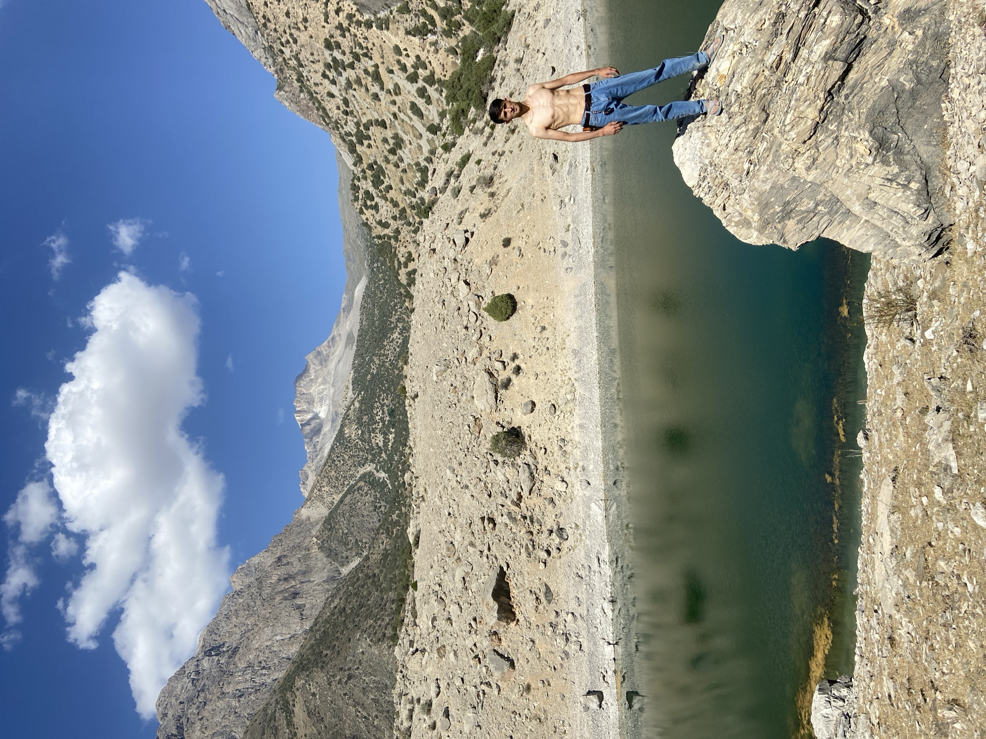 A young man with dark hair stands shirtless in blue jeans on a large, rocky outcropping overlooking a serene, green lake in the mountainous region of Ghazza, Tajikistan. The background features steep, arid mountainsides dotted with sparse vegetation, bathed in bright sunlight under a clear blue sky with scattered white clouds. The lake's surface is calm, reflecting the surrounding landscape and sky. The foreground consists of dry, rocky terrain leading to the water's edge. The man is positioned to the right of the frame, facing the camera with a relaxed stance.