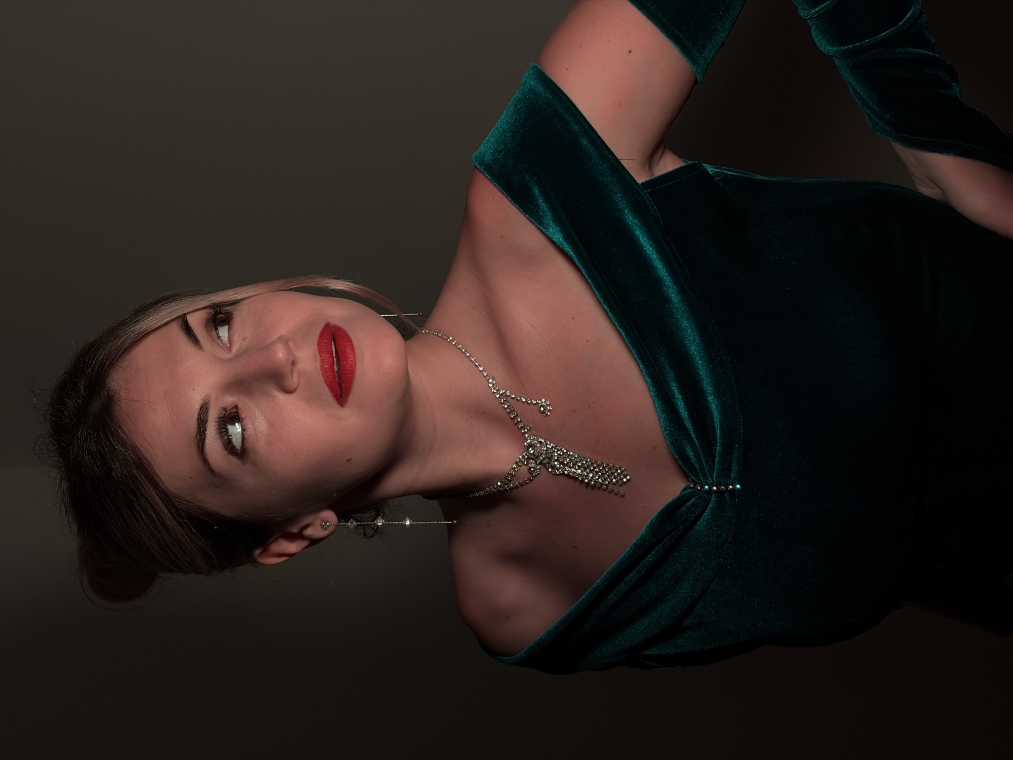 A woman with fair skin and blonde hair, wearing a dark green velvet dress and a silver necklace, is viewed from a low angle against a dark grey background. She has red lipstick and is wearing long, dangling earrings. The setting is dark and appears to be indoors. The image is rotated clockwise, making it appear as if the woman is leaning. Budva, Montenegro, is not evident in the image.