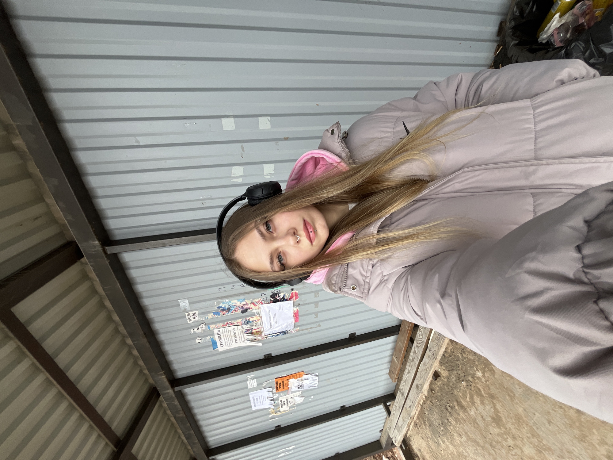 A young woman with long blonde hair is pictured in what appears to be an enclosed area constructed of corrugated metal. She wears headphones, a light purple puffer jacket with a pink hoodie underneath, and has facial piercings on her nose and lip. The woman looks towards the camera, tilting her head slightly. Various paper notices are stuck to the metal wall behind her. Near the top right of the frame, a trash bag is partially visible. The scene includes parts of wooden structures. Based on the surrounding context, the location is in the city of Фоки, Russia.