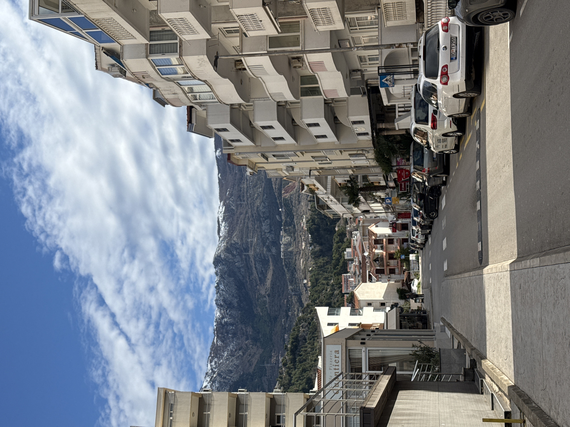The image depicts a street view in Budva, Montenegro. The scene is taken from an elevated position, likely from a building. The street is lined with buildings on both sides, with parked cars along the right side. In the distance, a large, snow-capped mountain is visible against a blue sky with clouds. A building with a sign "Pizzeria" is visible in the foreground. The street appears narrow and leads towards the mountain in the distance. There is no evident presence of people in this image. The focus is on the urban landscape juxtaposed with the mountainous backdrop.