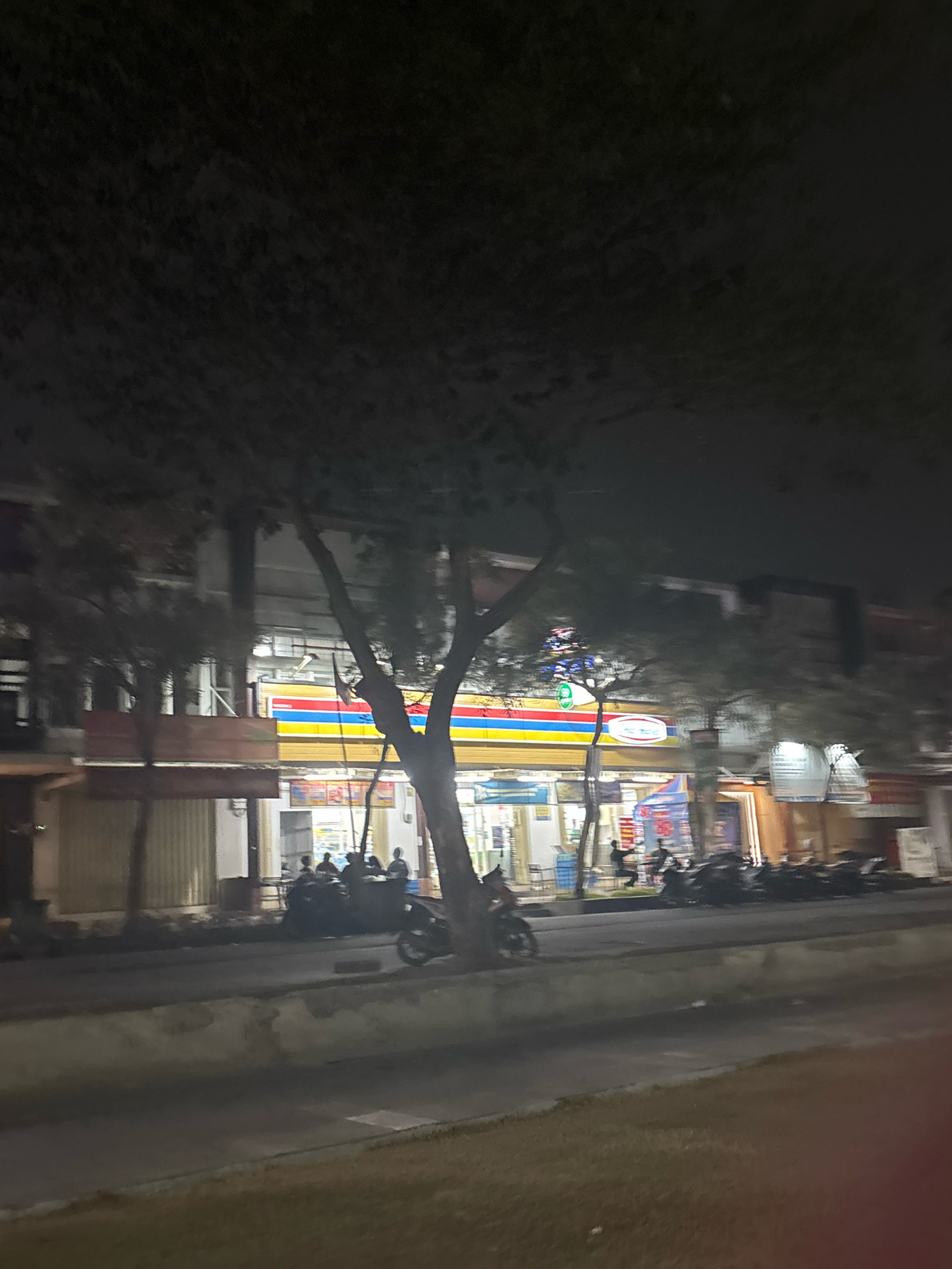 The image depicts a nighttime street scene in Telagamurni, Indonesia. A bright storefront with horizontal bands of yellow, blue, and red signage dominates the middle ground, suggesting a commercial establishment such as a convenience store. Several motorcycles are parked in front of the store and along the street. A group of individuals, silhouetted against the store's light, are gathered near the entrance. Trees with dense foliage partially obscure the view of the buildings and the sky. The street in the foreground appears to be paved, with a raised concrete median or curb. The overall ambiance is that of a busy urban area at night, illuminated by artificial lighting.