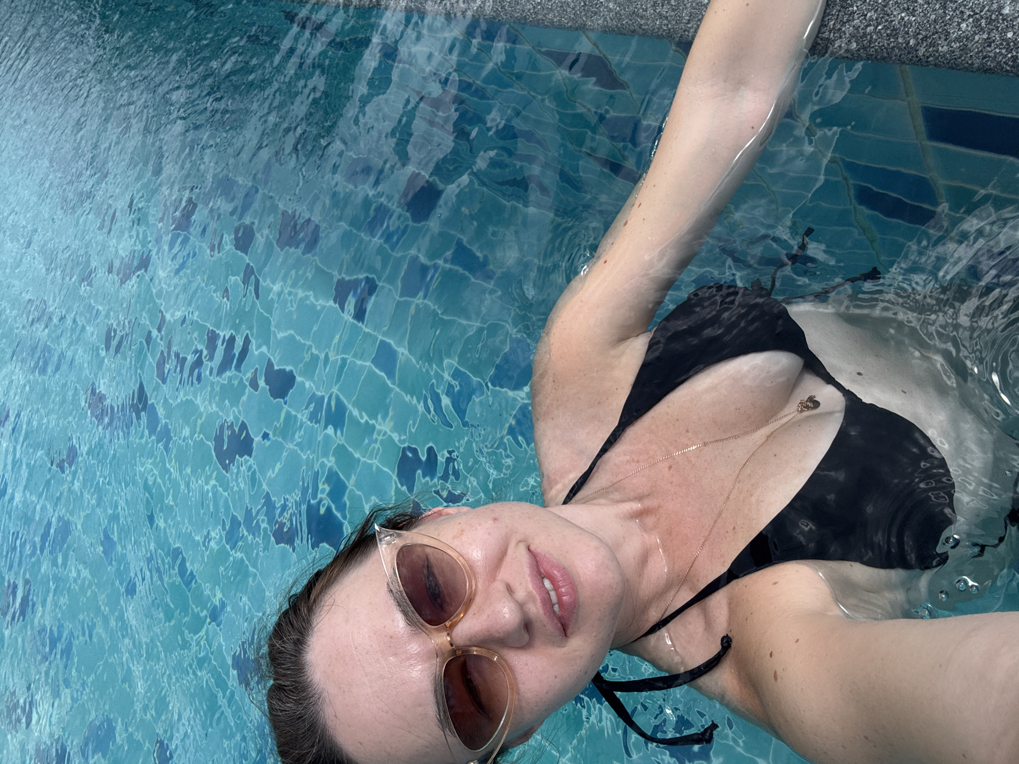 A person is floating on their back in a swimming pool. They are wearing a black bikini top and light-colored sunglasses. The pool has blue tiled walls and the water is clear and rippling. The person's head is tilted back, and they appear relaxed. The sunlight suggests it is daytime, and the setting appears to be a tropical location, possibly a resort, given the context of Baan Plai Laem, Thailand. There are no visible signs or text in the image.