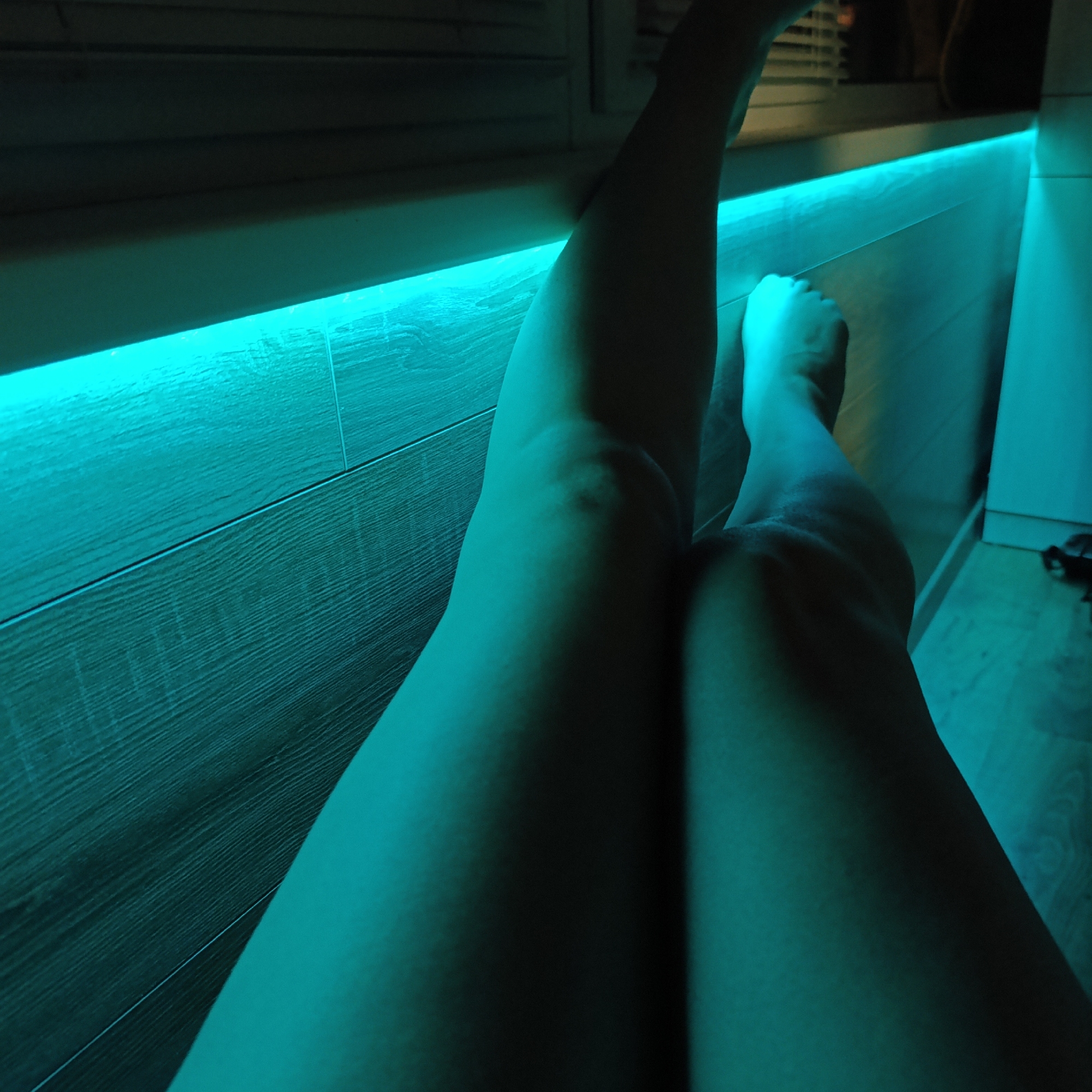 The image shows a pair of legs resting against a wall with blue LED lighting. The wall appears to be made of wood paneling. The feet are visible. The location in the city of Марковский, Russia is not discernible from the image.