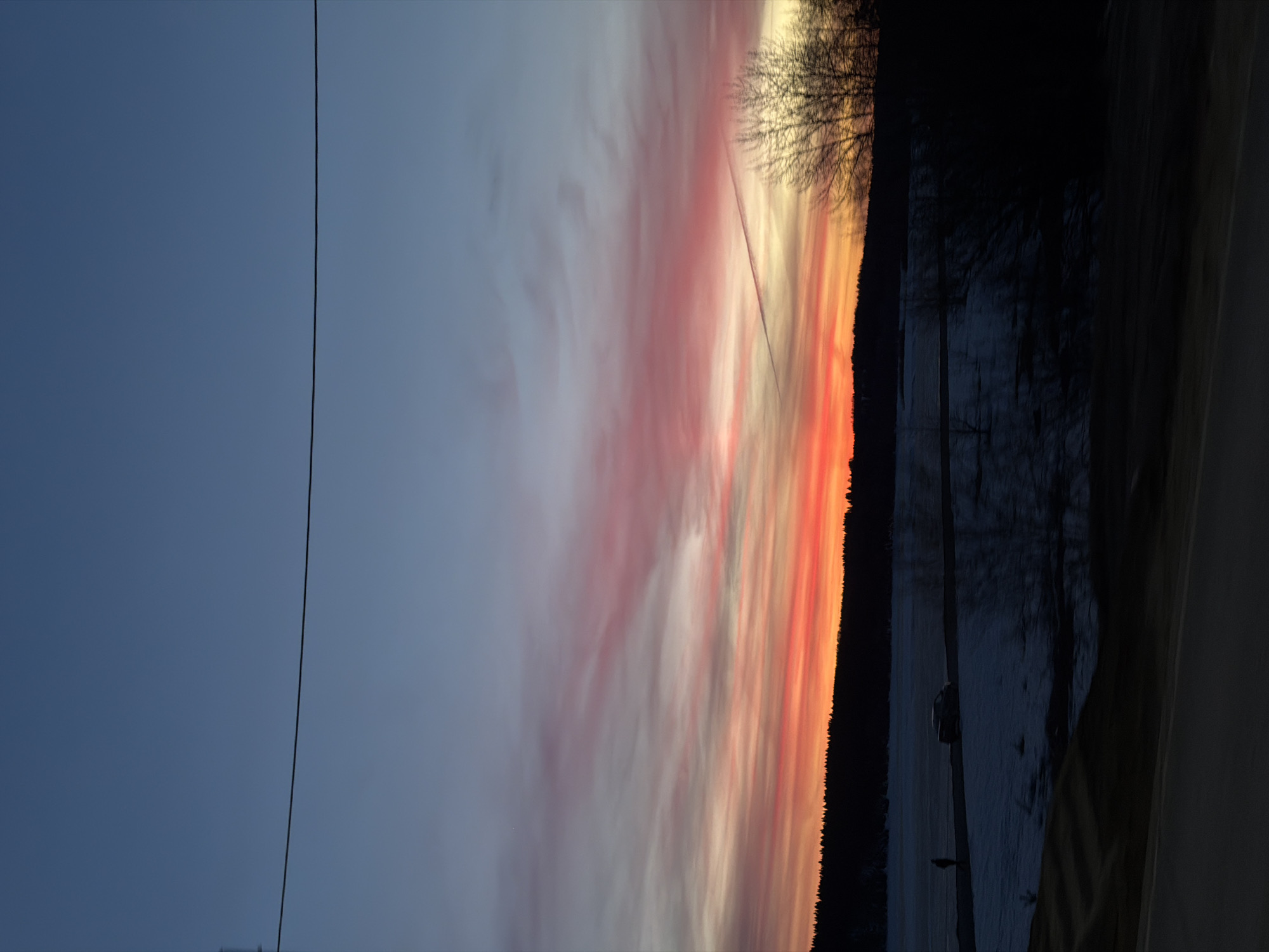 The image shows a colorful sunset with pink and orange hues. The sky fades from a dark blue on the left to vibrant colors toward the right, near a dark treeline. A thin, straight line, possibly a power line, runs vertically through the image on the left. On the right, beyond the treeline, there are hints of a body of water and some structures. The location is likely near a natural landscape feature in Usolye, Russia, given the context.
