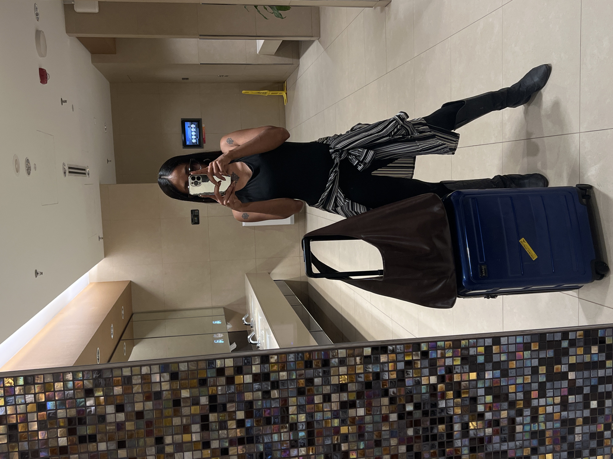A woman is taking a mirror selfie in a well-lit airport restroom. She is wearing a black tank top, black leggings, tall black boots, and a striped shirt tied around her waist. She is holding a smartphone in her right hand, pointed towards the mirror. Next to her, a blue rolling suitcase is topped with a dark brown hobo bag. The restroom features a long, integrated sink counter with multiple faucets, a tiled wall with colorful mosaic accents, and neutral-toned floor tiles. A digital display screen is visible on the back wall.