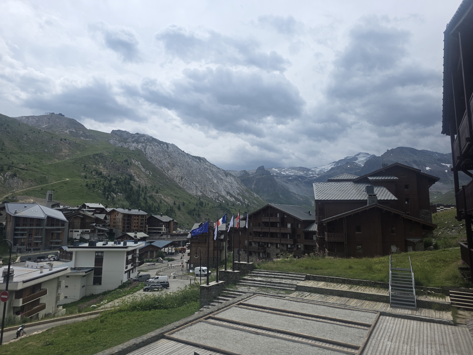 The image depicts a village scene in the French Alps, likely in Tignes, France, given the architecture and mountain landscape typical of ski resorts. The overcast sky suggests it is not a summer day. Several buildings, constructed with dark wood and grey roofs, dominate the mid-ground. These appear to be chalets or apartment buildings, characteristic of mountain resorts. In the foreground, there is a wooden deck with a bocce ball court, and a metal staircase leads up to a grassy area.
Several flags, including the European Union flag, the French flag, and other national or organizational flags, are displayed on poles near the buildings. Cars are parked on a road that winds through the village. The background is defined by steep, green mountains, with patches of rock and some snow visible on higher peaks, suggesting a high-altitude location.
The overall impression is a quiet, somewhat cloudy day in a mountain resort town, with a mix of residential or tourist accommodations and recreational facilities. No people are clearly visible performing any specific action, though the presence of cars implies inhabitants or visitors. The architecture and the flags point to a European, likely French, setting.