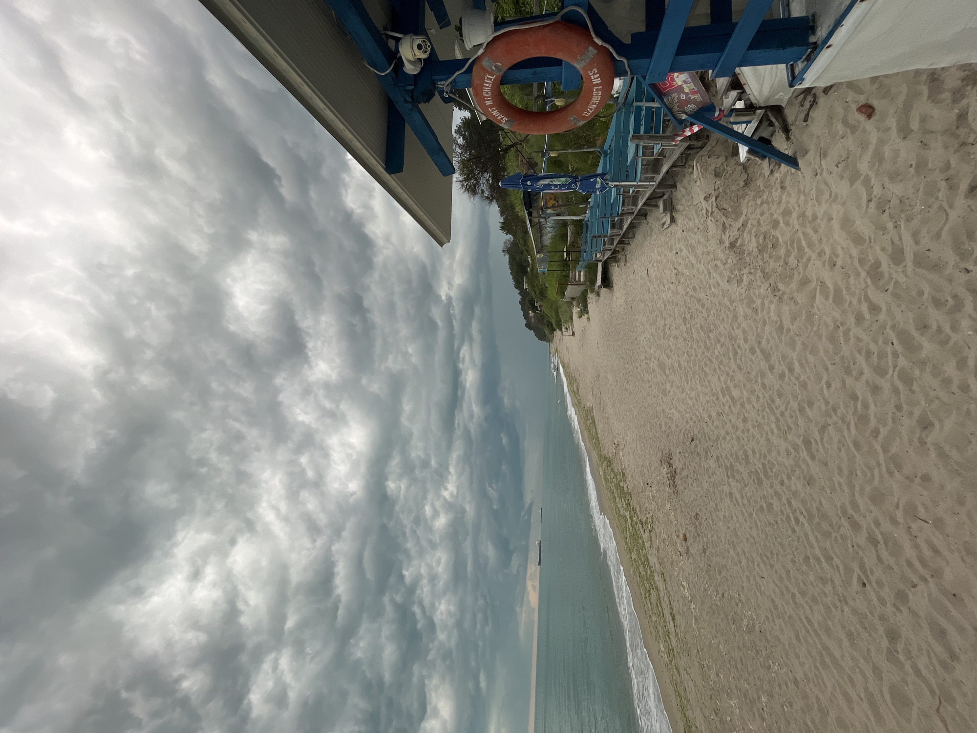 The image displays a coastal scene, likely a beach, under a heavily overcast sky. The photograph is oriented vertically, showing a long stretch of sandy beach and the ocean extending into the distance on the right, and a prominent blue wooden structure with an overhang on the left.
No people are visible in the frame. Key objects include an orange lifebuoy hanging from the blue wooden structure, which also features what appears to be a security camera mounted near the top left. The lifebuoy has visible text: "SAINT MICHAEL" and "SAN LORENZO." The blue structure itself consists of wooden beams and planks, suggesting a beach facility such as a bar, changing room, or lifeguard stand. Faintly visible behind the structure are hints of greenery, possibly trees or bushes, and another blue beach umbrella.
The sandy beach shows numerous footprints, indicating recent activity, and a thin line of green vegetation or seaweed along the water's edge. The ocean appears calm with small waves breaking on the shore, and several ships or cargo vessels are visible on the horizon. The sky is filled with thick, textured grey clouds, suggesting an overcast day, possibly before or after rain, lending a muted and calm atmosphere to the scene. The overall lighting is soft and diffused. The location is Varna, Bulgaria.