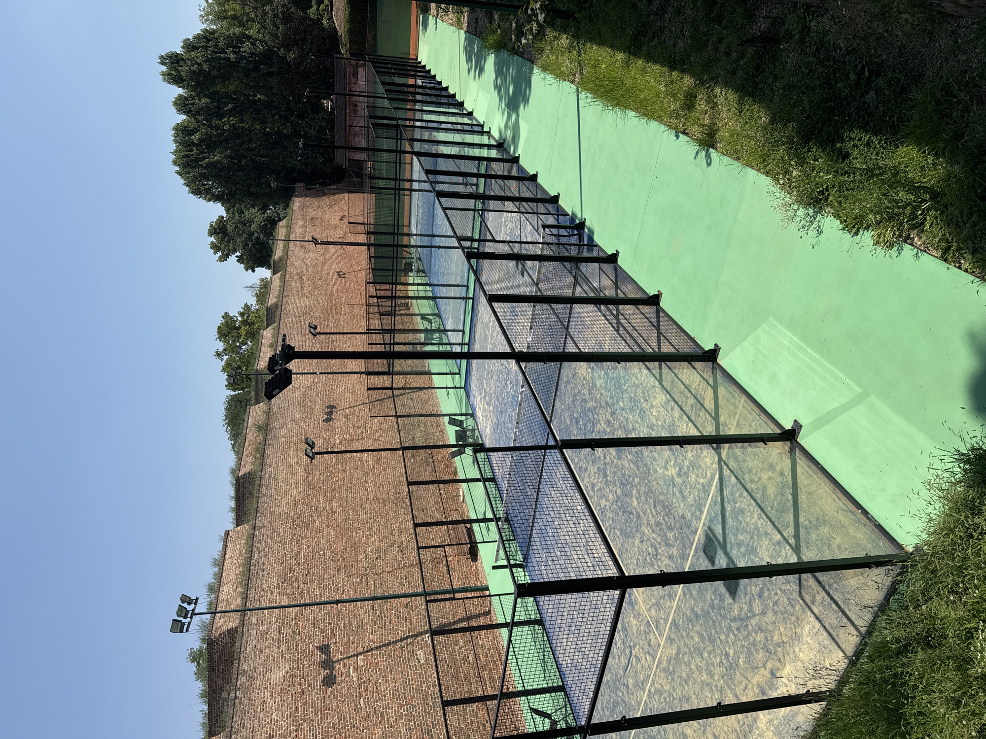The image shows multiple outdoor paddle tennis courts with bright green ground surfaces and glass walls. The courts are enclosed by black metal fencing. In the background, there is a weathered brick wall that appears to be part of a historic fortification. Several tall, dark metal light poles with multiple lamps are visible, illuminating the courts. Trees and vegetation are present behind the brick wall. The sky is clear and blue, indicating a sunny day. The scene is likely located in an urban setting, possibly within the Kalemegdan Fortress in Belgrade, Serbia, given the architectural style of the brick wall. No people are visible in the image.