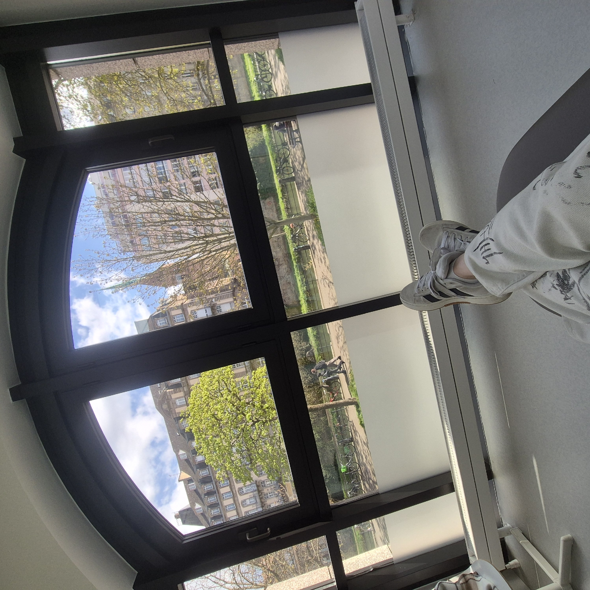 A person is sitting by a large, arched window. The window frame is black, and through the window, buildings and trees can be seen. Outside, there is a river with people walking along the bank. Bicycles are parked along the path. A portion of blinds are drawn in front of part of the window. The person is wearing white sneakers and light-colored jeans.
Based on the architecture, particularly the church spire and building style, the location outside the window appears to be in Strasbourg, France. The river is likely the Ill.
