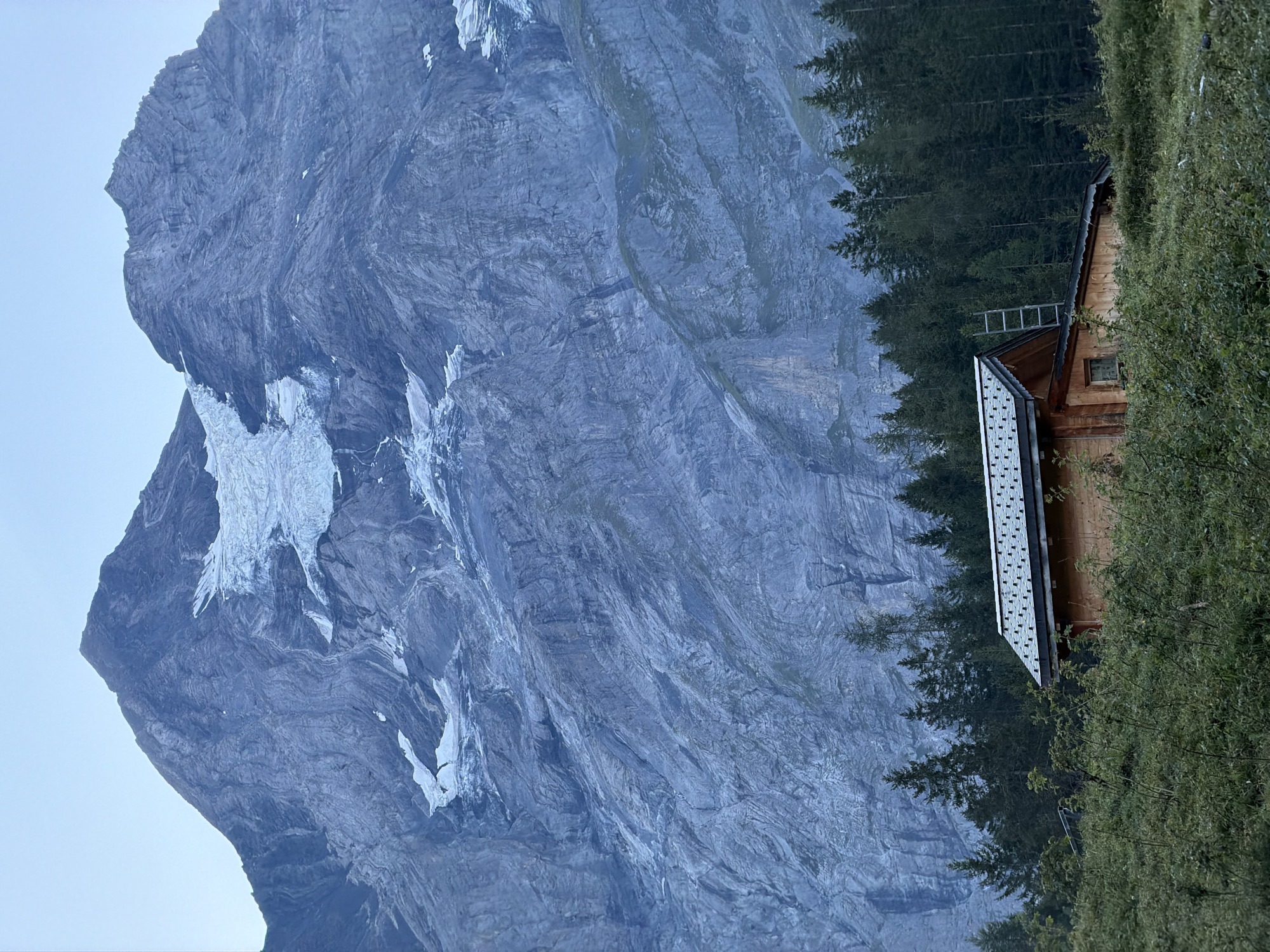 A wooden cabin with a gray, shingled roof is situated on a grassy slope in front of a dense forest. The cabin has a small window and a ladder leaning against its side. Behind the forest, a massive, rocky mountain with a snow-covered peak and glacier dominates the background. The sky above is a pale, hazy blue. The scene appears to be located in the Swiss Alps, likely near Kandersteg, Switzerland, given the characteristic mountain landscape.