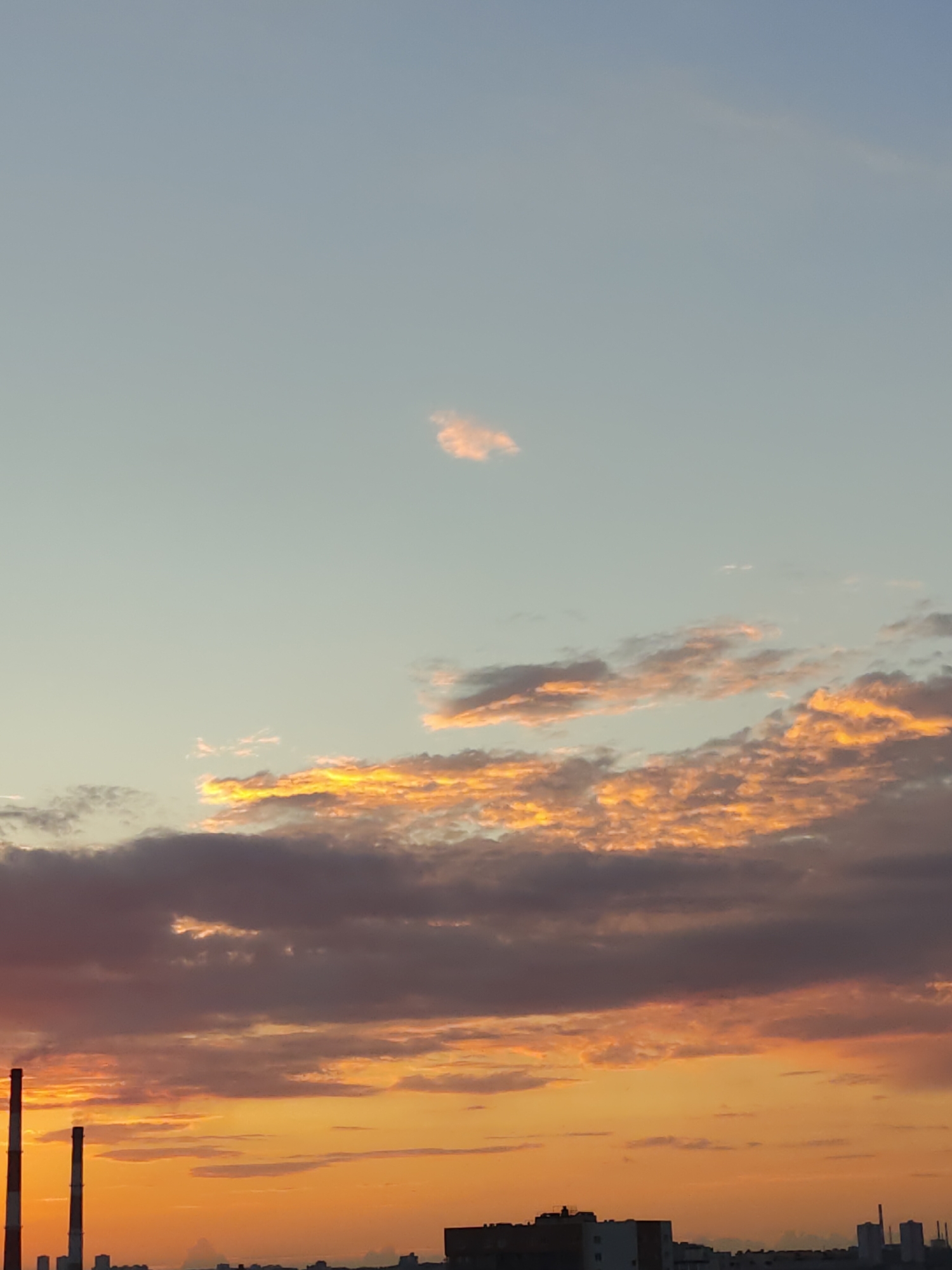 This is an outdoor, full shot, eye-level photograph of a cloudy sky above an industrial cityscape at sunset. The sky transitions from a pale blue at the top to a vibrant orange at the horizon. The clouds are tinged with yellow and orange, with darker gray clouds in the foreground. Two tall, red and white striped smokestacks are prominent on the left side of the frame, suggesting an industrial area. In the distance, a silhouetted cityscape with various buildings and another smokestack can be seen. The overall scene captures a dramatic sunset over an industrial part of an unknown city.