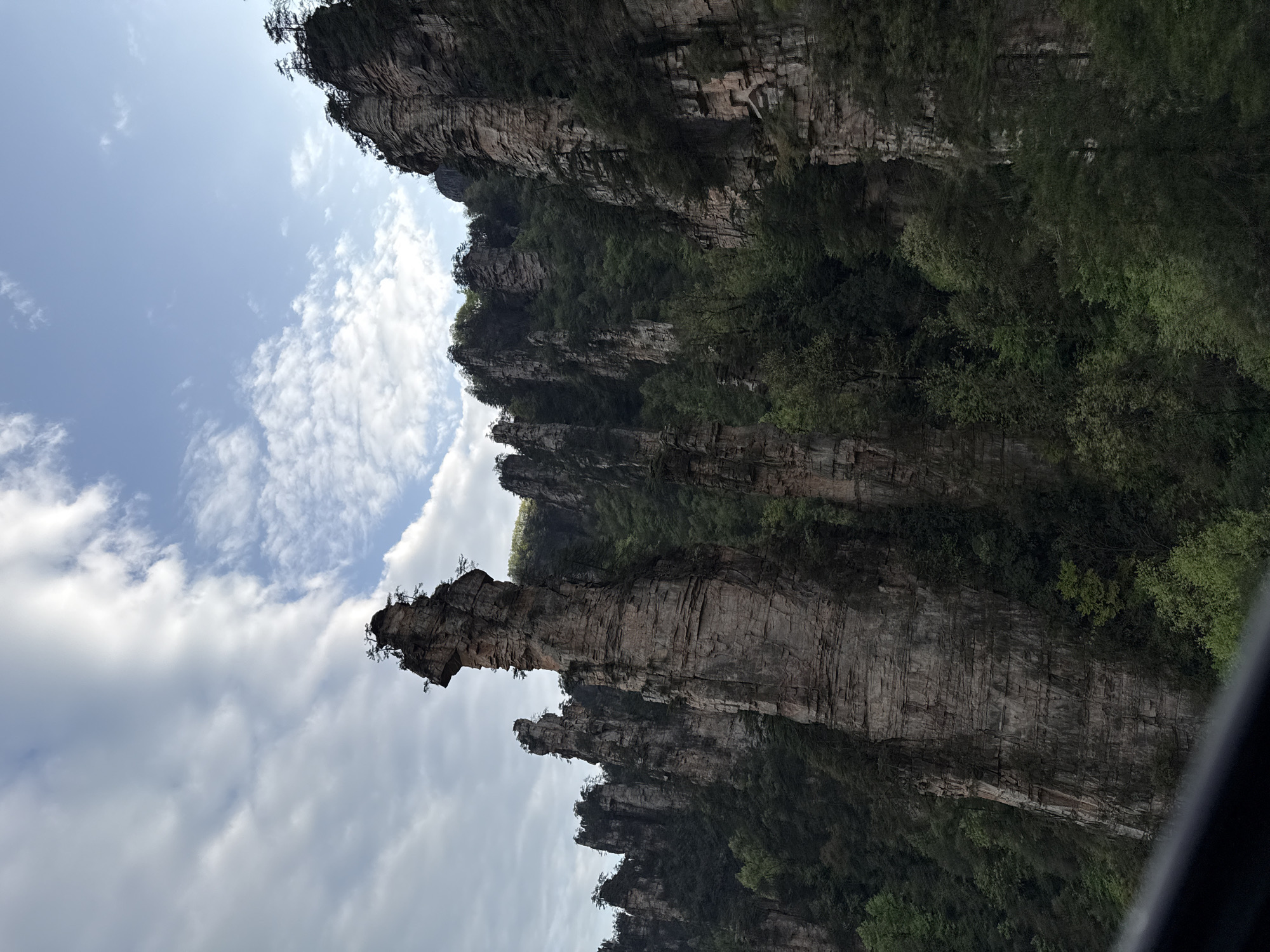 The image depicts a mountainous landscape in Wulingyuan, China. Towering sandstone pillars with vegetation cling to their sides. The sky is visible with light blue hues interspersed with white clouds. There are no people visible in the scene.