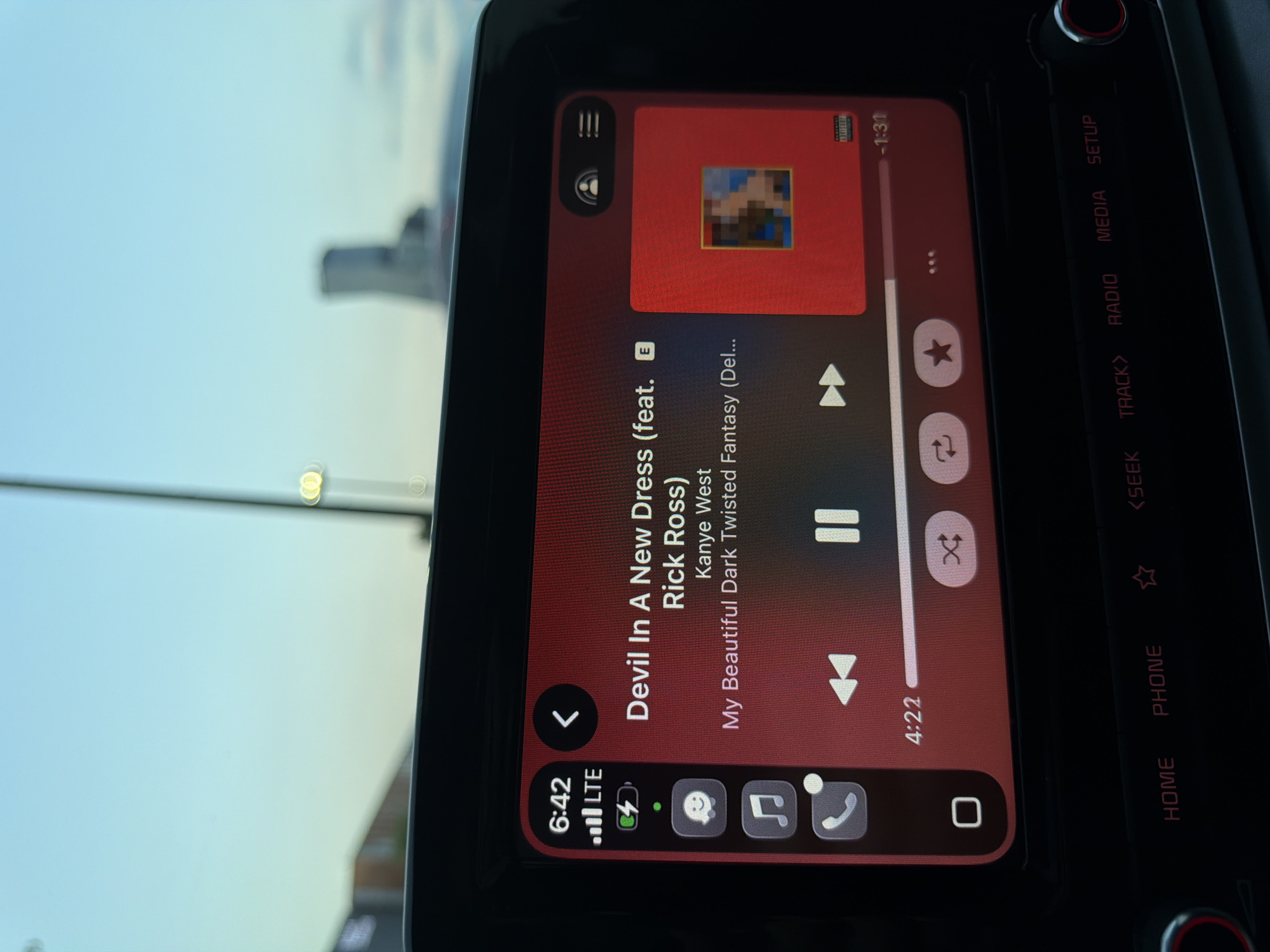 This media file presents a close-up view of a car's infotainment system, rotated 90 degrees counter-clockwise. The screen prominently displays a music player interface with a dark red theme. The song "Devil In A New Dress (feat. Rick Ross)" by Kanye West from the album "My Beautiful Dark Twisted Fantasy" is shown as playing. The album cover art is visible but pixelated or censored. The current playback time indicates 4:22 into the song, with -1:31 remaining.
At the bottom left of the screen, a status bar shows the time as 6:42, "LTE" cellular signal strength, and a charging battery icon. Additional icons for phone calls and other applications are present. To the right of the screen, physical buttons for car functions are visible, labeled "HOME", "PHONE", "<SEEK", "TRACK>", "RADIO", "MEDIA", and "SETUP".
Through the car window, a clear, light blue sky can be seen, suggesting either early morning or late afternoon/dusk. A blurred object, possibly a distant building or another vehicle, is faintly visible in the background. The scene takes place inside a vehicle, possibly in Puebla, Mexico.