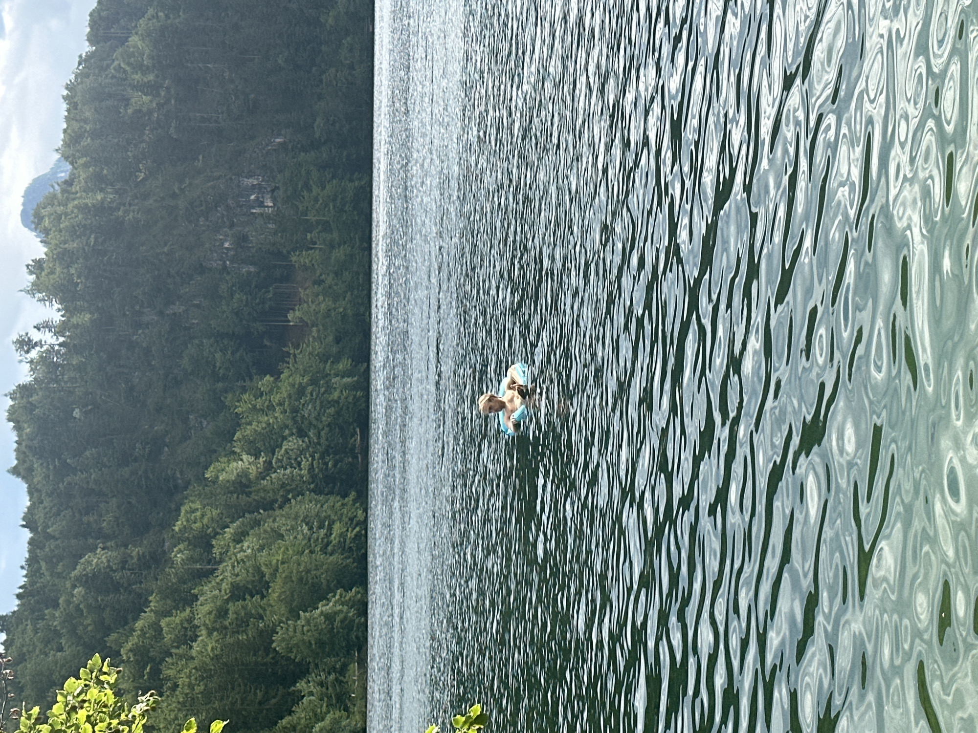 A person with blonde hair is floating in a blue inflatable ring in a body of water. The water is dark and appears wavy. In the background, a dense forest covers a hillside, with a peak visible above the trees. The scene is set in Kiefersfelden, Germany.