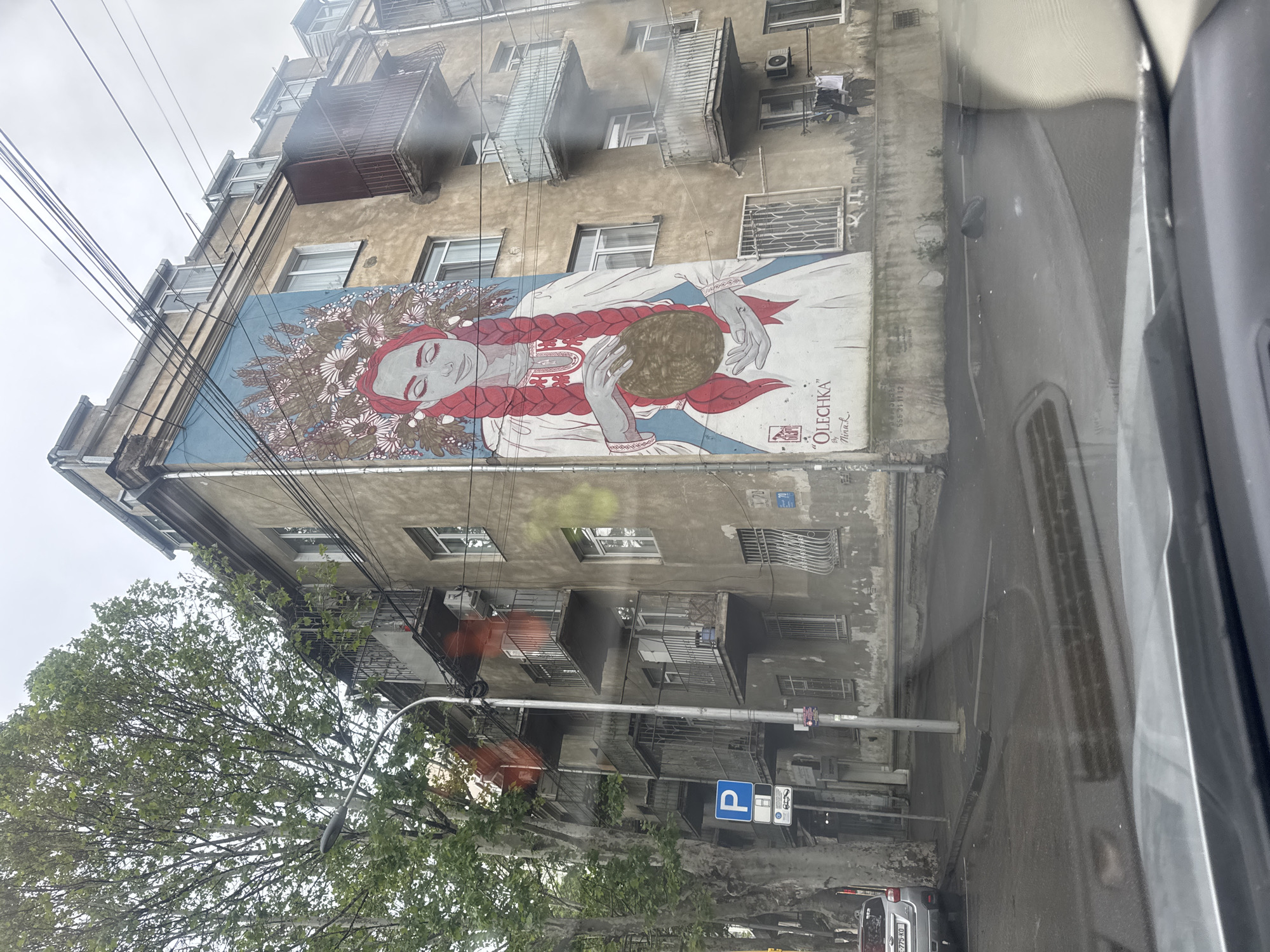 The image depicts a multi-story building in Tbilisi, Georgia, featuring a large mural of a woman with a flower crown and red braids, holding a round object. The mural is located on the side of the building. The building has small balconies and visible utility lines. A tree with green leaves is partially visible in the lower left corner. A street with a parking sign is in the foreground. The photo appears to have been taken from inside a vehicle, with part of the car's interior visible on the right side of the image.