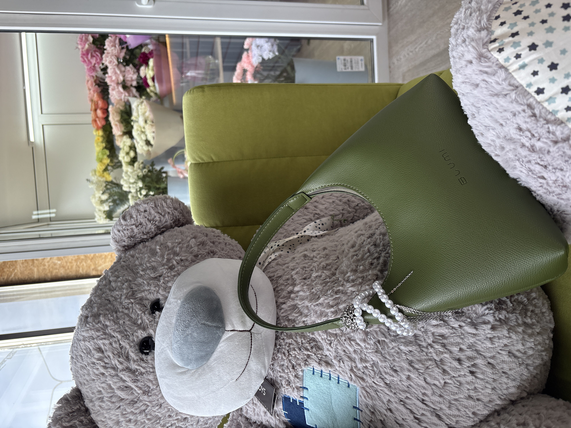 The image shows a gray stuffed teddy bear with a white snout and a blue patch on its body. Resting on the bear is a green leather handbag. The bear and bag are on a green upholstered chair. In the background, visible through a window, is a display of artificial flowers and foliage. The flowers are in shades of pink, white, and green. The location is Krasnoyarsk, Russia.