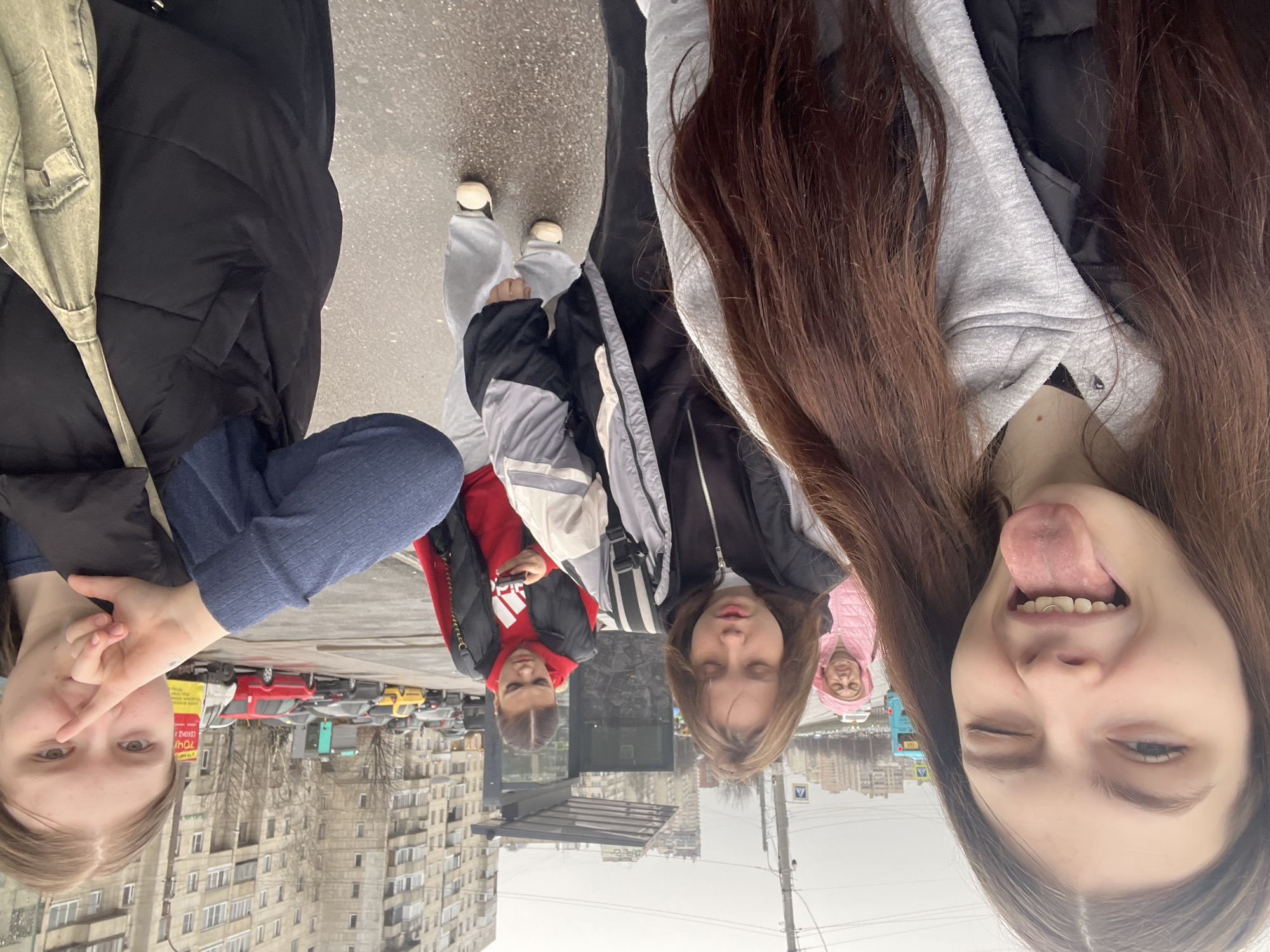 The image captures a group of five young people posing for a selfie in what appears to be a city street. The individuals are arranged closely, with parts of their bodies and faces visible in the frame. The foreground is dominated by the faces of the people, some showing playful expressions.
In the background, buildings of Saint Petersburg, Russia can be seen, indicating an urban setting. There are several cars parked along the street and typical urban street furniture can be seen, such as street lights and signs.
