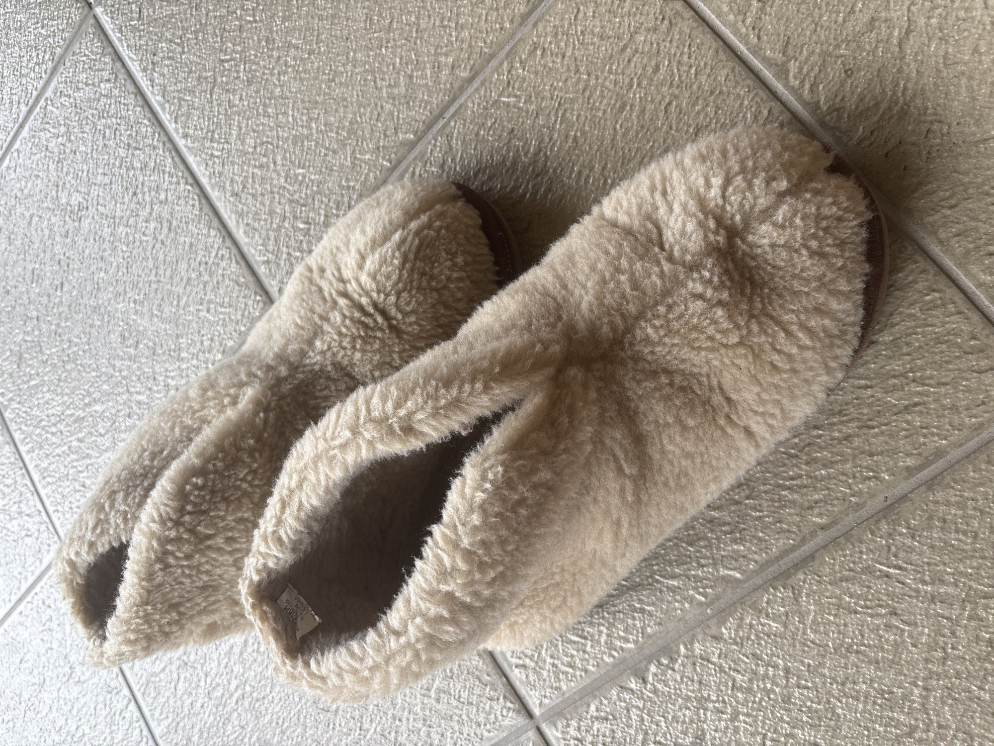 A pair of light brown, fuzzy, slipper-style shoes are shown from a top-down perspective on a tiled floor. The shoes are made of a plush, sheepskin-like material. The left shoe is in the foreground and the right shoe is slightly behind and to the right. The shoes are resting on a light-colored tiled surface with visible grout lines. No people are present. The scene appears to be indoors, likely in a residential setting within the Пятрышкаўскі сельскі Савет, Belarus.